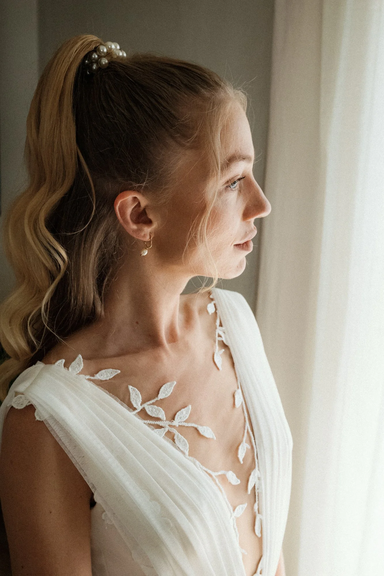 Junge Frau in einem weißen Kleid blickt nach rechts vor einem Fenster, trägt eine Perlen-Haarspange und einen Ohrring mit Perle.