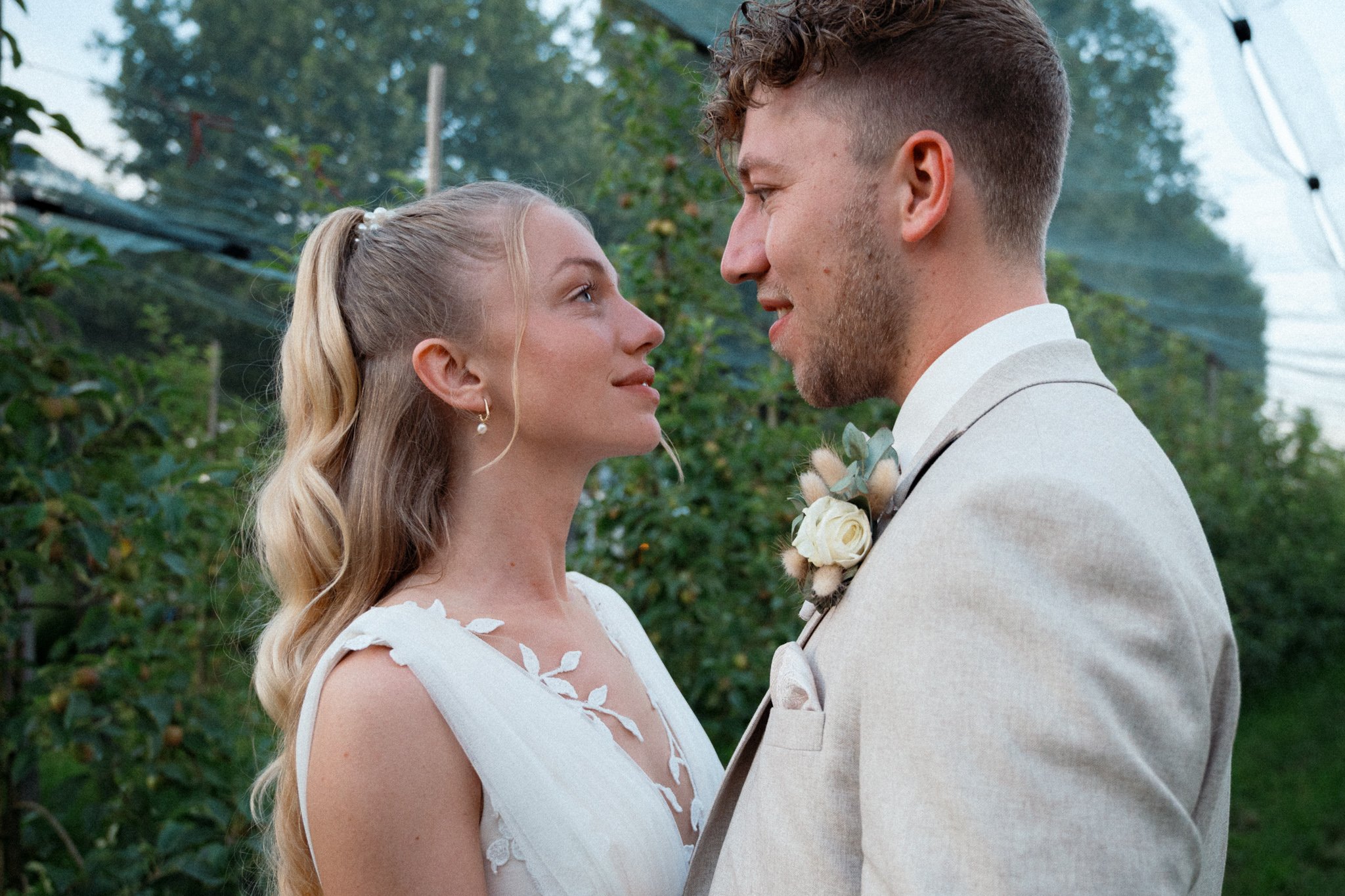 Ein Brautpaar schaut sich liebevoll in die Augen. Die Frau trägt ein weißes Kleid mit Blumenmuster, der Mann trägt einen hellen Anzug. Sie stehen im Freien vor grünen Büschen und Bäumen.
