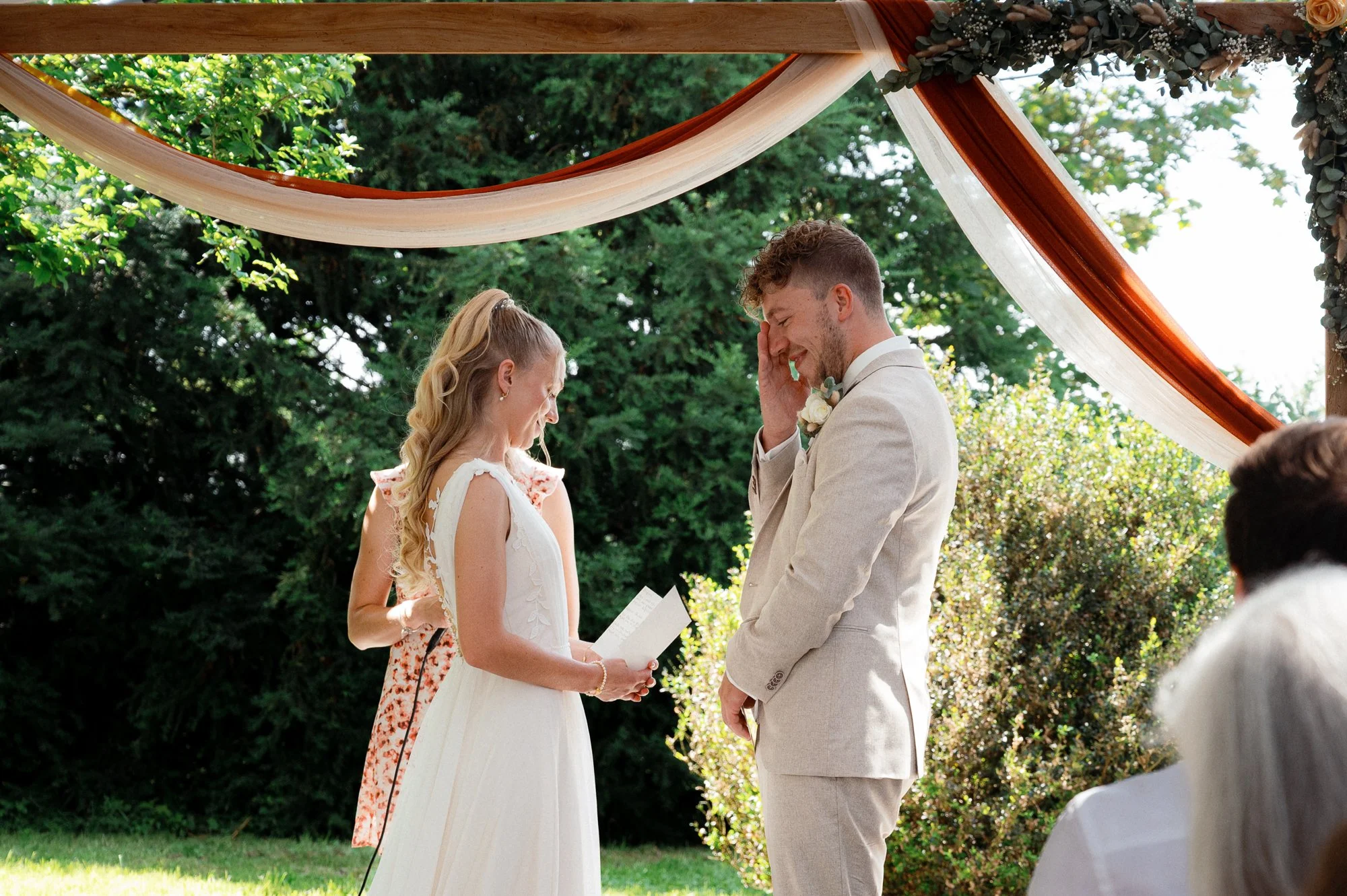Ein Brautpaar bei einer standesamtlichen Trauung im Freien mit einem Zeugen. Die Frau trägt ein weißes Hochzeitskleid, der Mann einen hellen Anzug. Sie stehen unter einem dekorierten Baldachin mit bunten Stoffen, umgeben von grünen Bäumen.