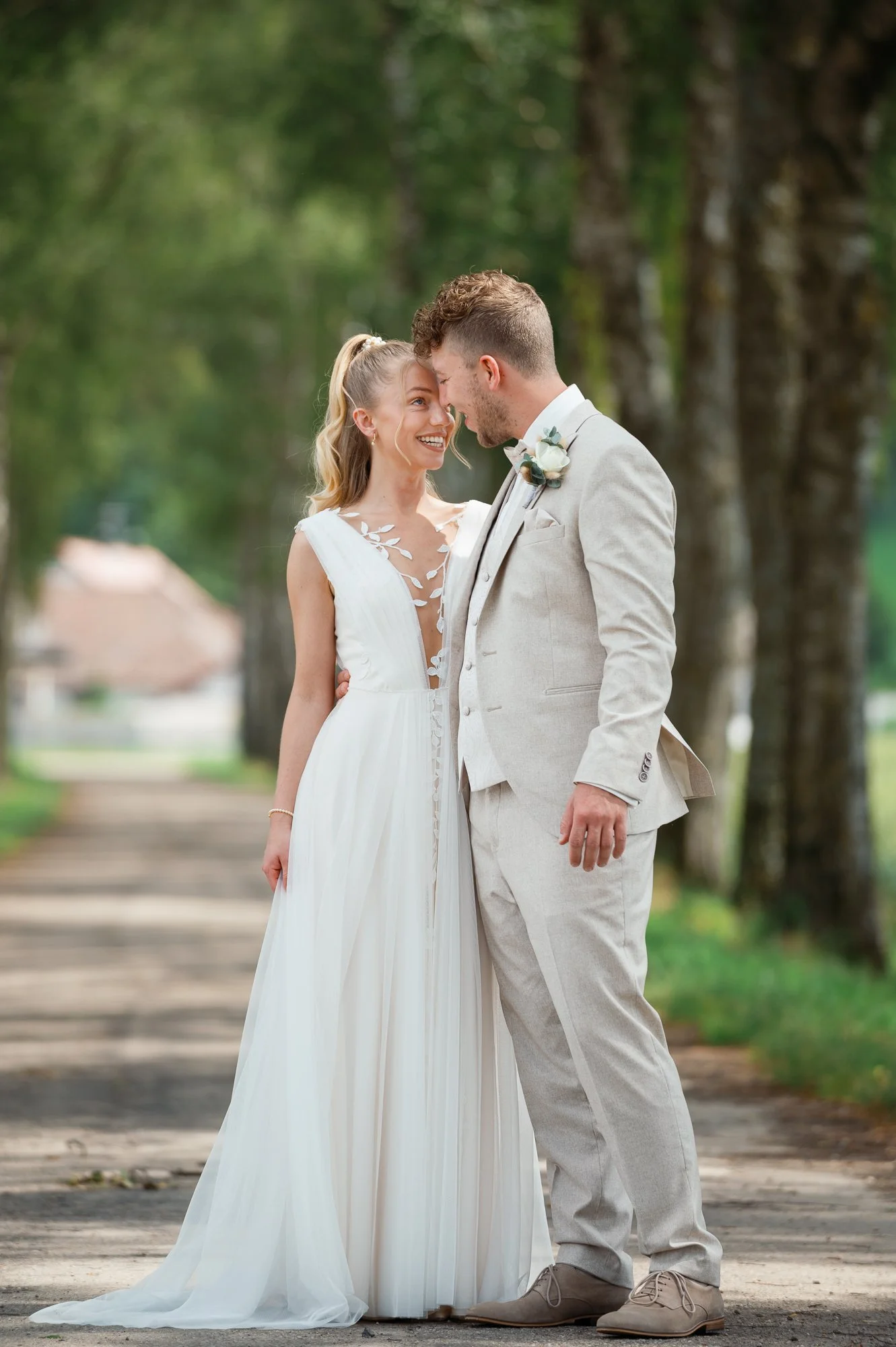 Ein Brautpaar steht eng beieinander im Freien, die Frau trägt ein elegantes weißes Hochzeitskleid und der Mann trägt einen hellbeigen Anzug, beide schauen sich liebevoll an, im Hintergrund sind Bäume und eine Straße.