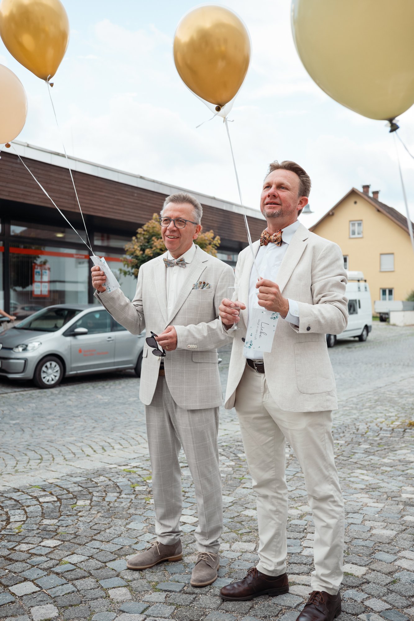 Zwei Männer in hellen Anzügen mit Krawatten stehen auf einem gepflasterten Platz und halten goldene und weiße Ballons an Schnüren. Im Hintergrund sind moderne und traditionelle Häuser. Es wirkt wie eine Feier oder ein besonderer Anlass.