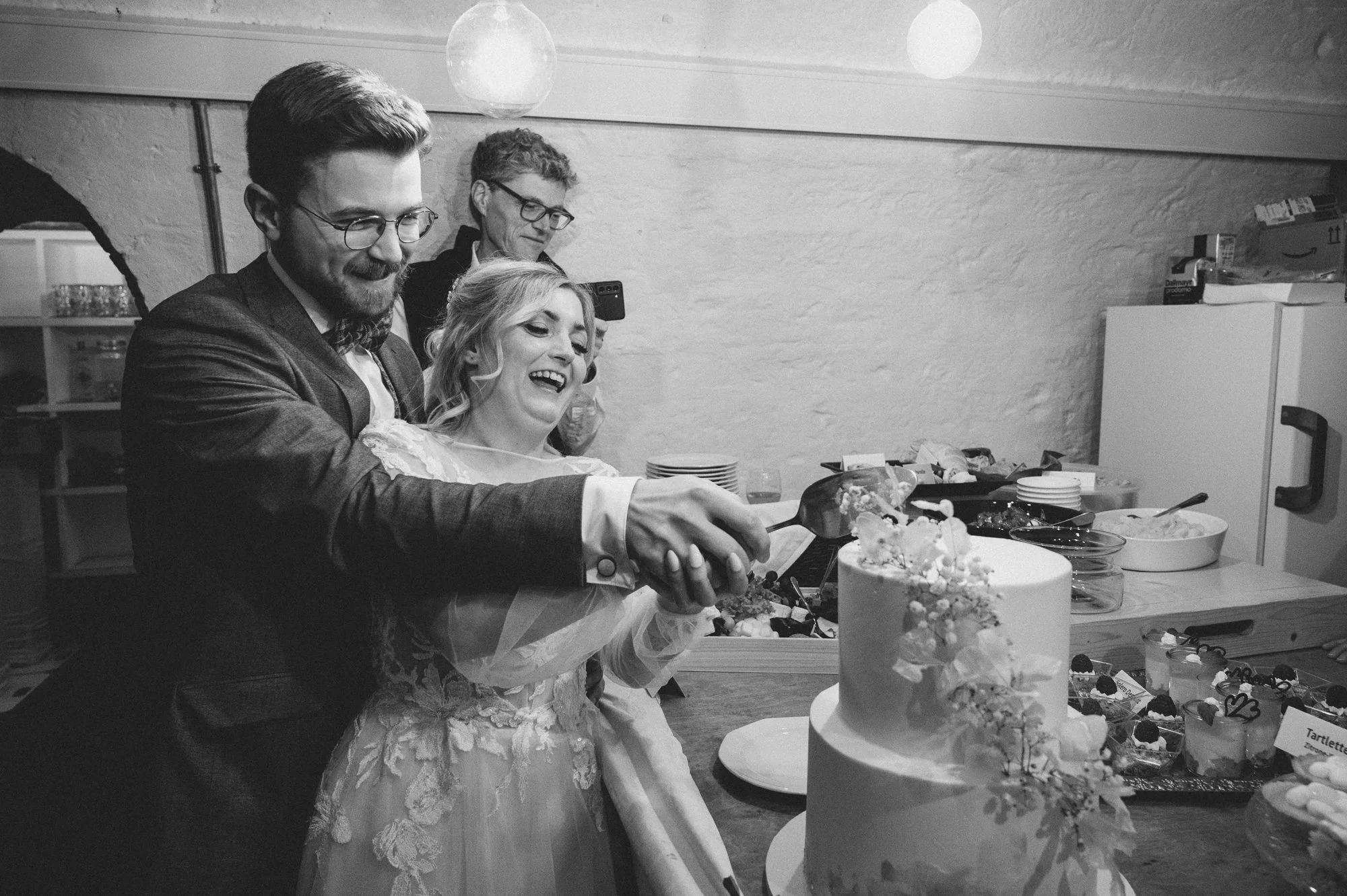 Drei Menschen, inklusive einer Frau in einem weißen Hochzeitskleid, schneiden eine Torte bei einer Feier. Es sind Teller und andere Desserts auf dem Tisch.