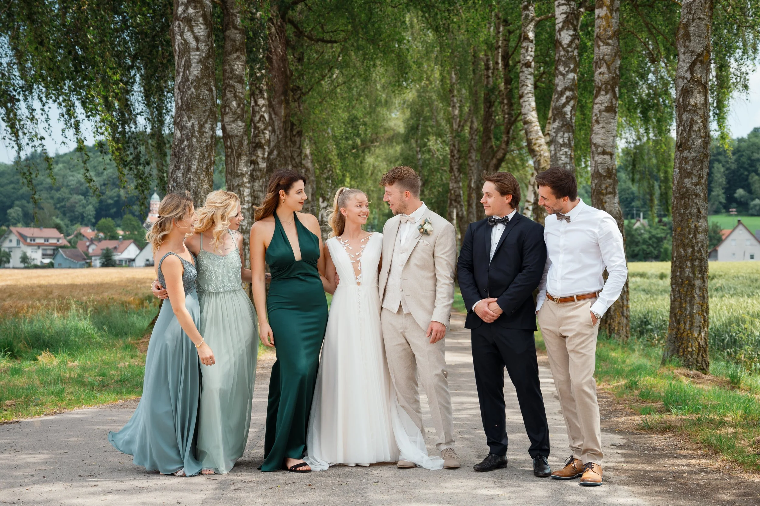 Eine Gruppe von sechs Menschen, bestehend aus sechs Frauen und zwei Männern, in formeller Kleidung, stehen in einer Zeile unter Bäumen in einer ländlichen Umgebung und sprechen miteinander bei einer Hochzeit im Freien.