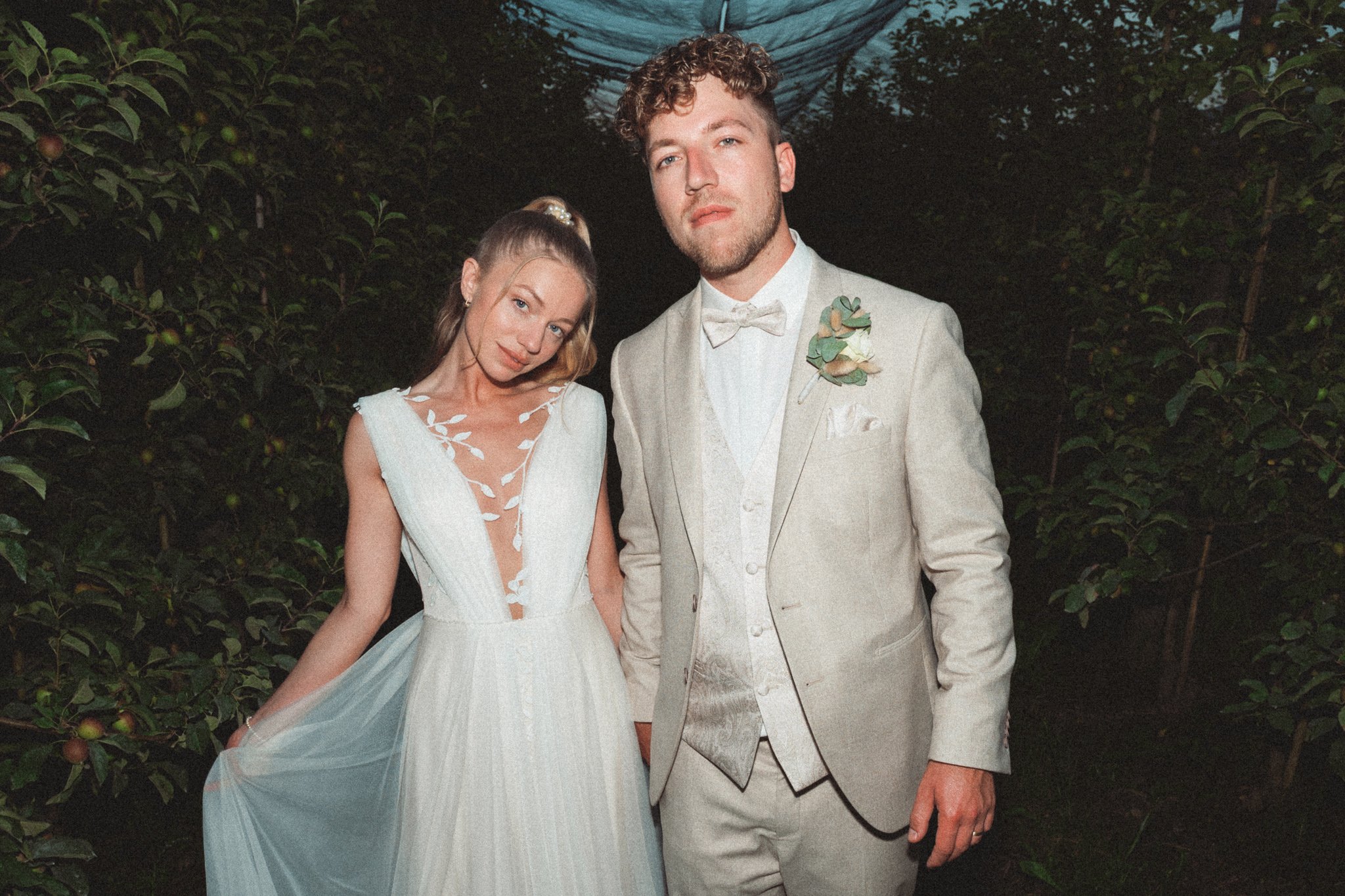Ein Paar bei einer Hochzeit, das in einem Garten steht. Die Frau trägt ein weißes Brautkleid mit Spitzen und eine Tüllschleppe, der Mann trägt einen hellgrauen Anzug mit weißem Hemd und Fliege.