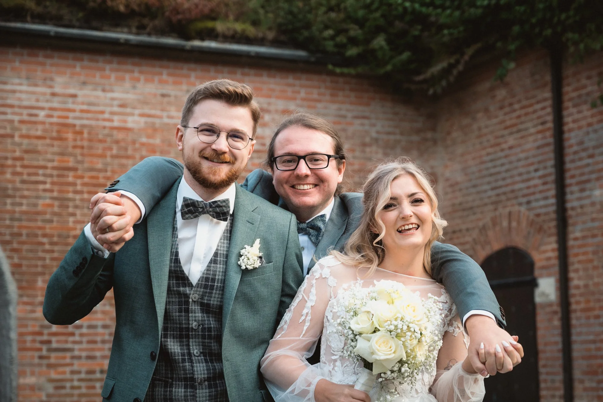 Drei Menschen bei einer Hochzeit, ein Mann im grauen Anzug und eine Frau in einem Hochzeitskleid, die alle lächeln und zusammenstehen.