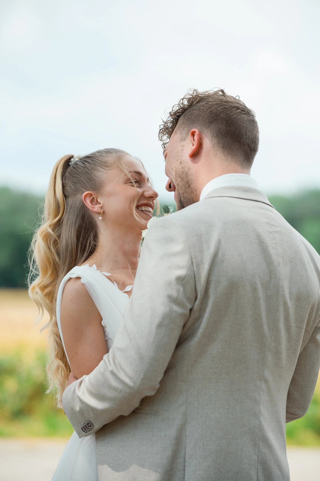 Ein Paar bei einer Hochzeit im Freien, umarmend, lächelnd, im Sommer mit bewölktem Himmel, im Hintergrund grüne Bäume und eine Wiese.