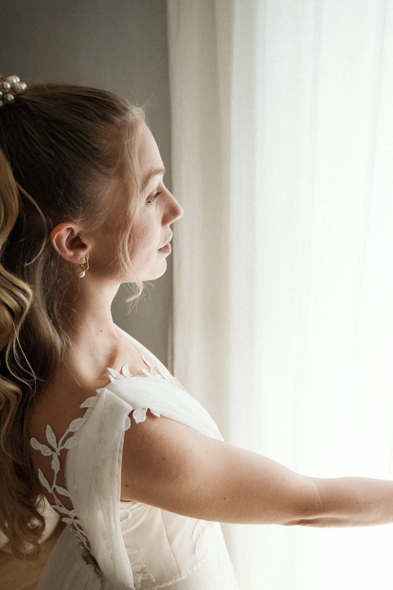 Nahaufnahme einer Frau im weißen Kleid, die neben einem Fenster steht und nach draußen schaut.