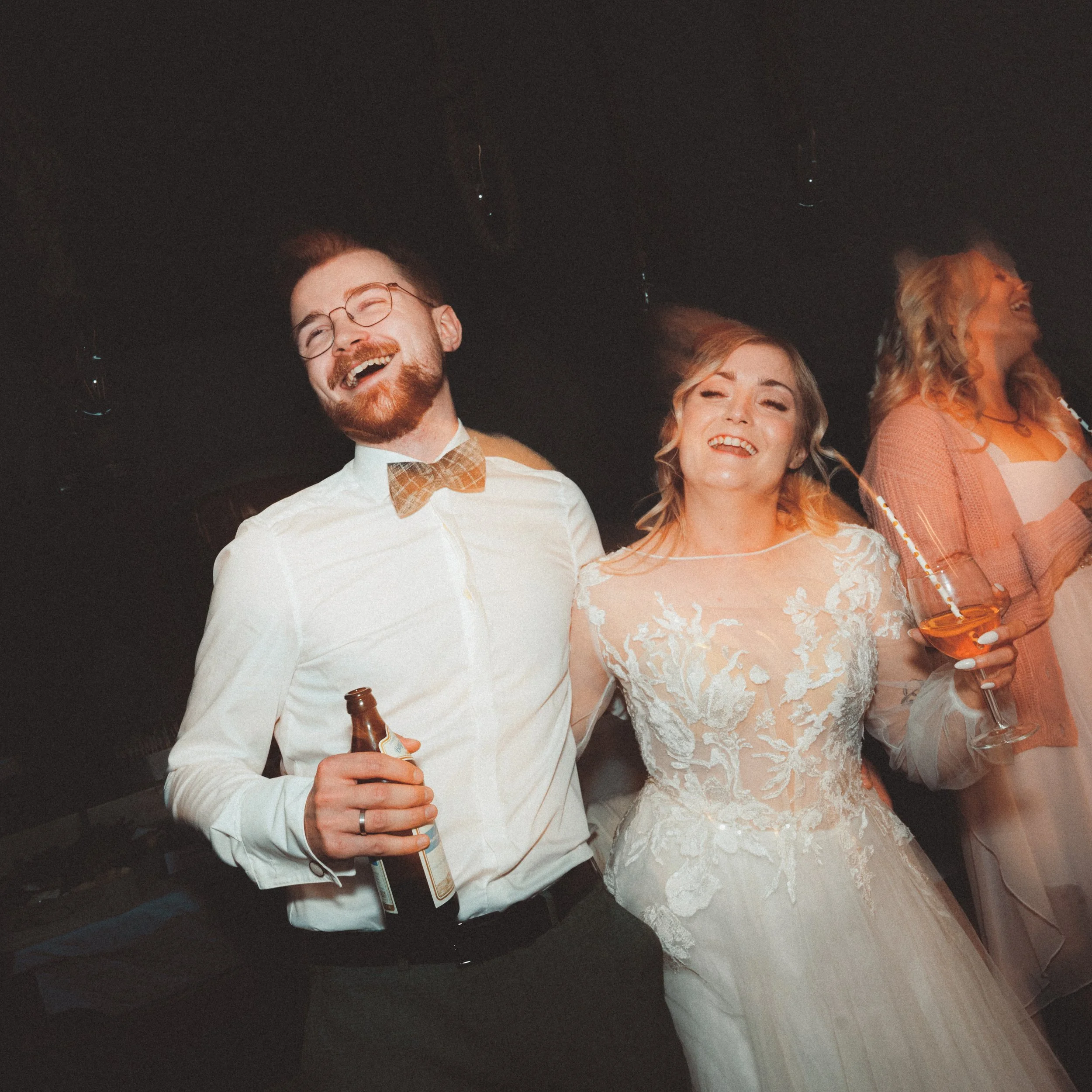 Menschen lachen und feiern auf einer Party, trinken Bier, einige tragen Brillen, im Hintergrund dunkler Raum.