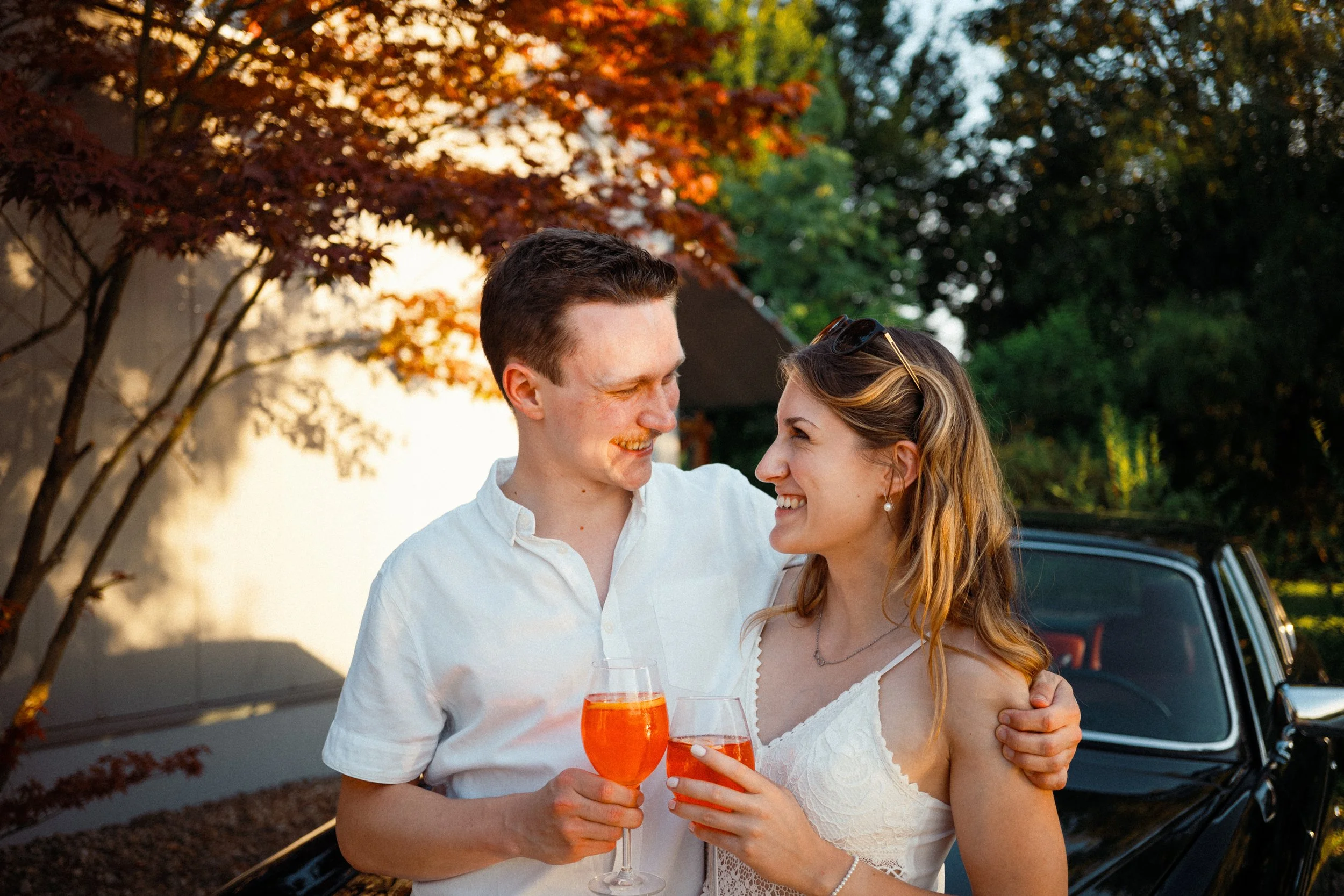 Ein Mann und eine Frau, die sich umarmen, halten Gläser mit orangefarbenem Getränk bei Sonnenuntergang im Freien, mit Bäumen mit roten und grünen Blättern im Hintergrund.