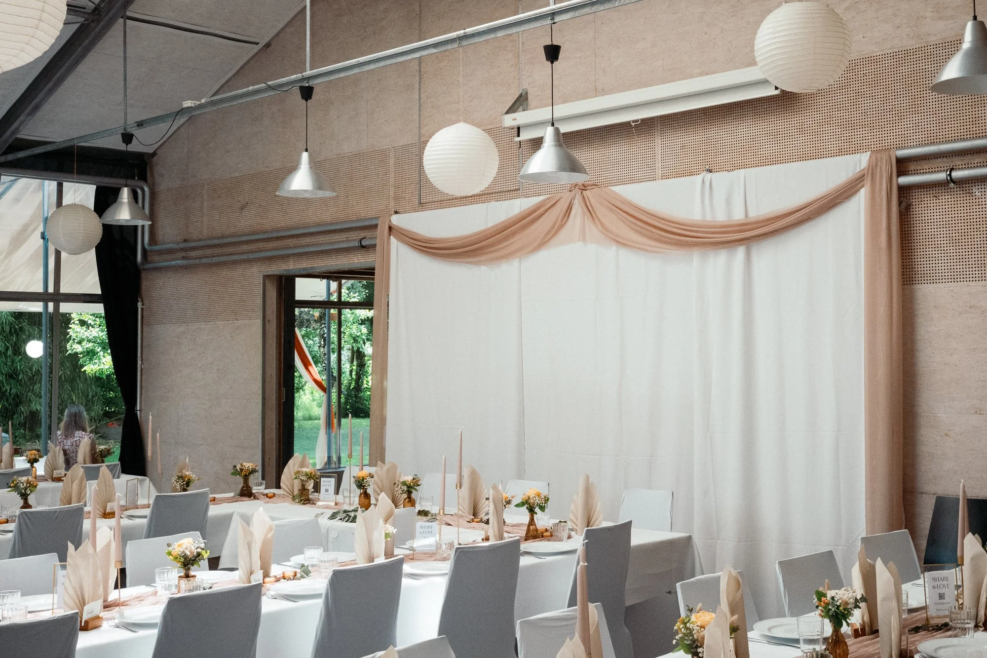Innenraum eines festlich gedeckten Raumes mit Tafel, weißen Stühlen, Blumen und Tischtuch, in einem großen Fenster sind Bäume zu sehen, während weiße Papierlaternen und metallene Lampen von der Decke hängen.