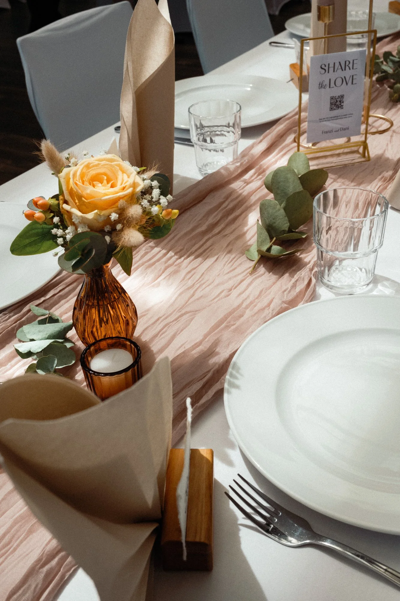 Ein festlich gedeckter Tisch mit weißem Geschirr, einer rosa Tischdecke, einem Blumenarrangement in einem braunen Glas, Kerzen und einem Namensschild, das zum Teilen der Liebe auffordert.