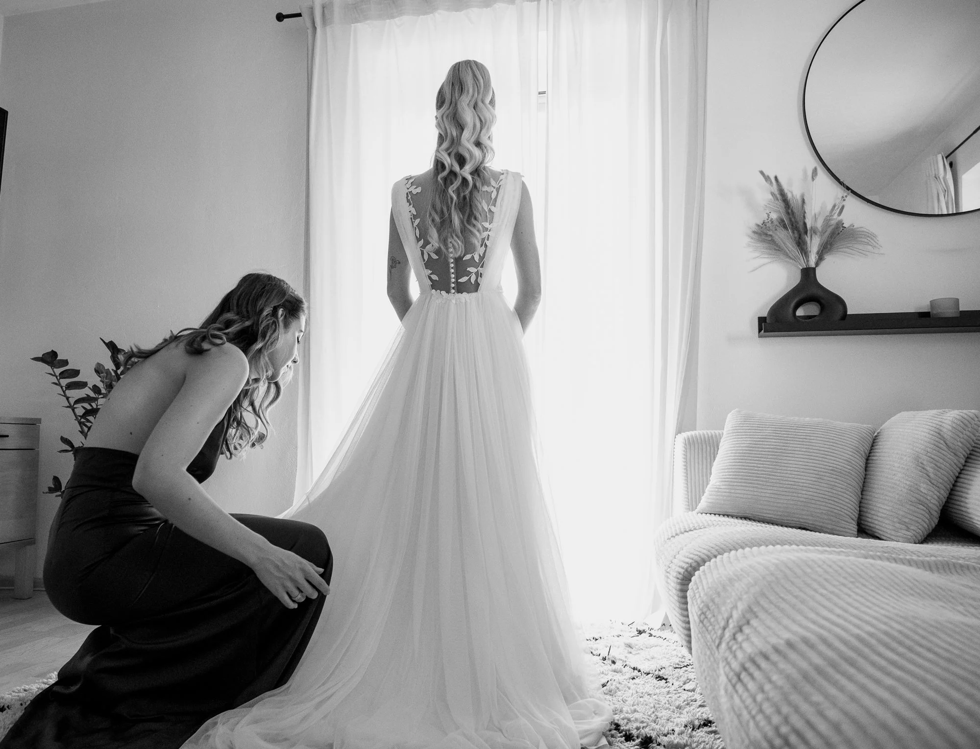 Zwei Frauen bereiten eine Braut vor, die im Schlafzimmer auf ihre Hochzeit wartet. Die Braut steht am Fenster in einem weißen Kleid, während eine Frau vor ihr sitzt, vermutlich zur Unterstützung oder Beratung.