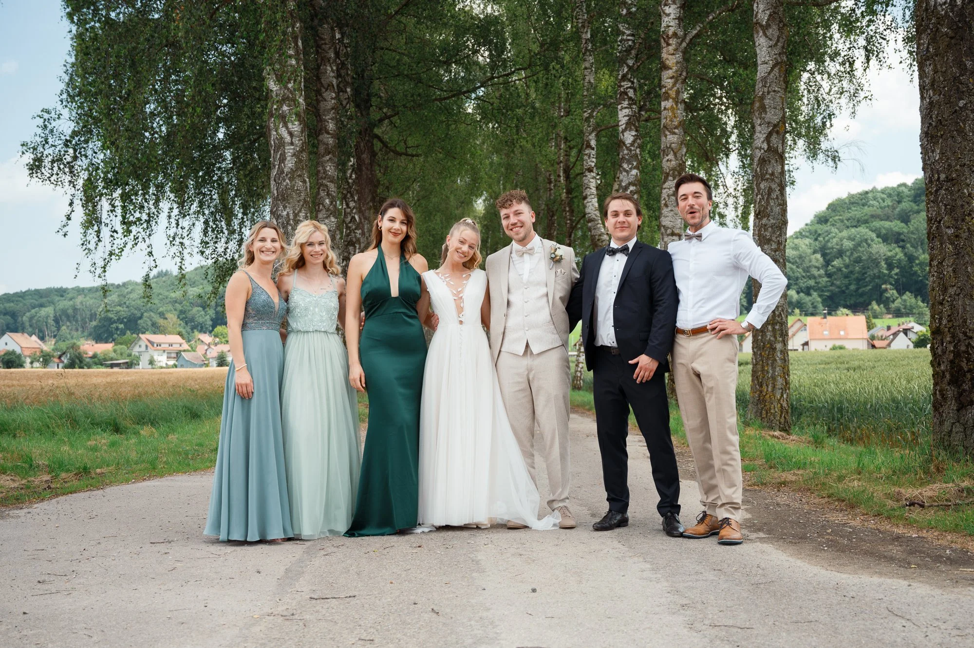 Eine Gruppe von sieben jungen Erwachsenen steht nebeneinander im Freien auf einem Weg, umgeben von Bäumen und einem kleinen Dorf im Hintergrund. Es scheint, dass sie an einer Hochzeit teilnehmen, da die beiden Männer Anzüge und die Frauen elegante Kl