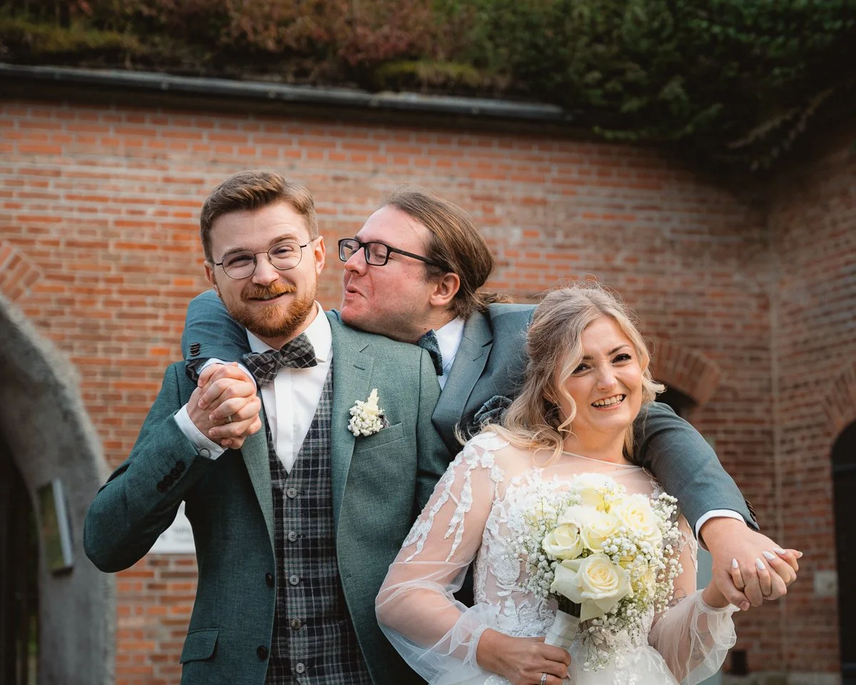 Eine Hochzeitsfeier draußen vor einer roten Ziegelmauer mit drei lachenden Menschen: Hochzeitspaar und ein junger Mann, der einem der Männer einen Kuss auf die Wange gibt.