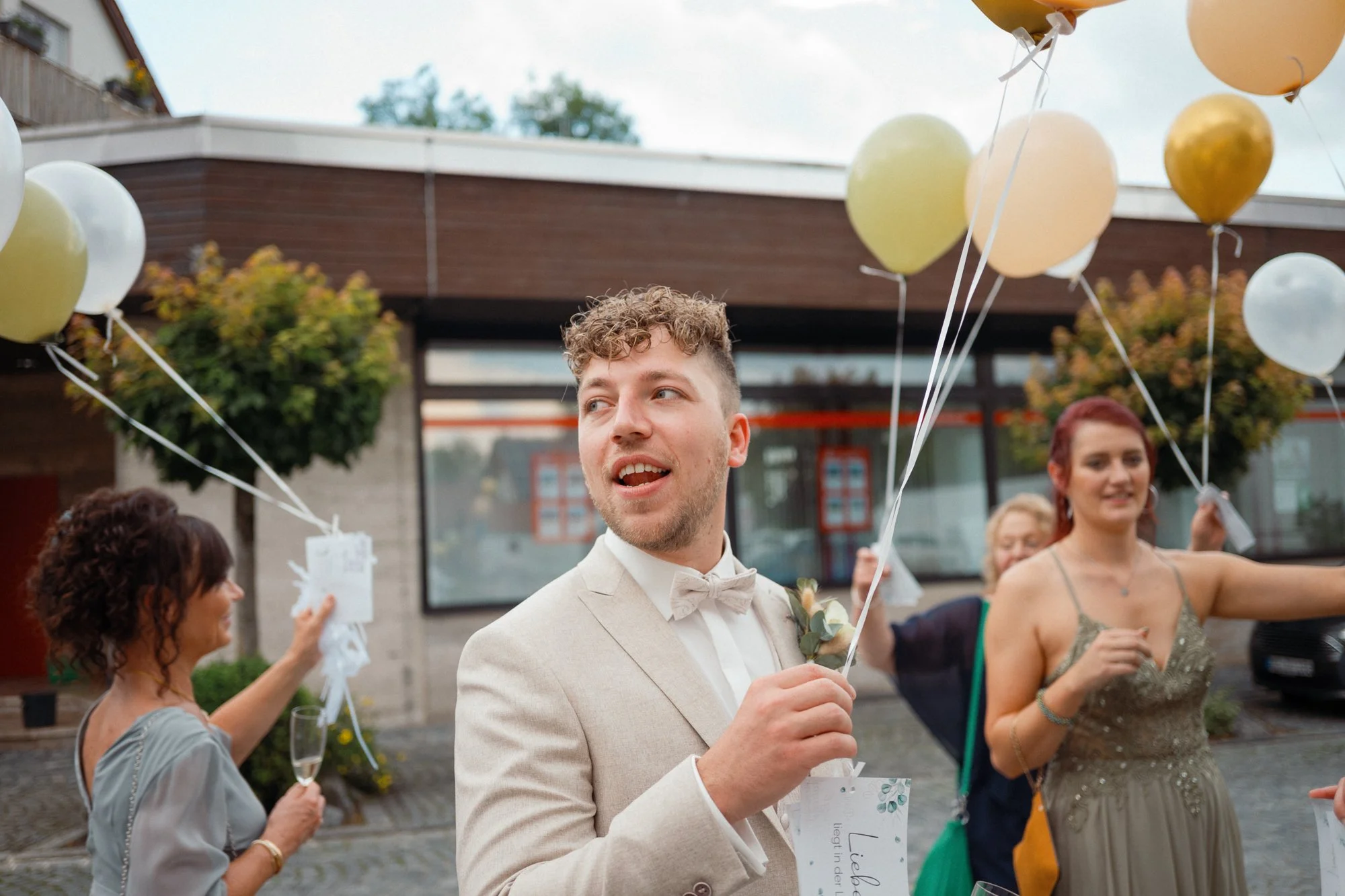 Junge Mann in Anzug mit Fliegen beim Feiern, umgeben von Frauen, die Ballons halten, bei einer outdoor Hochzeitsfeier