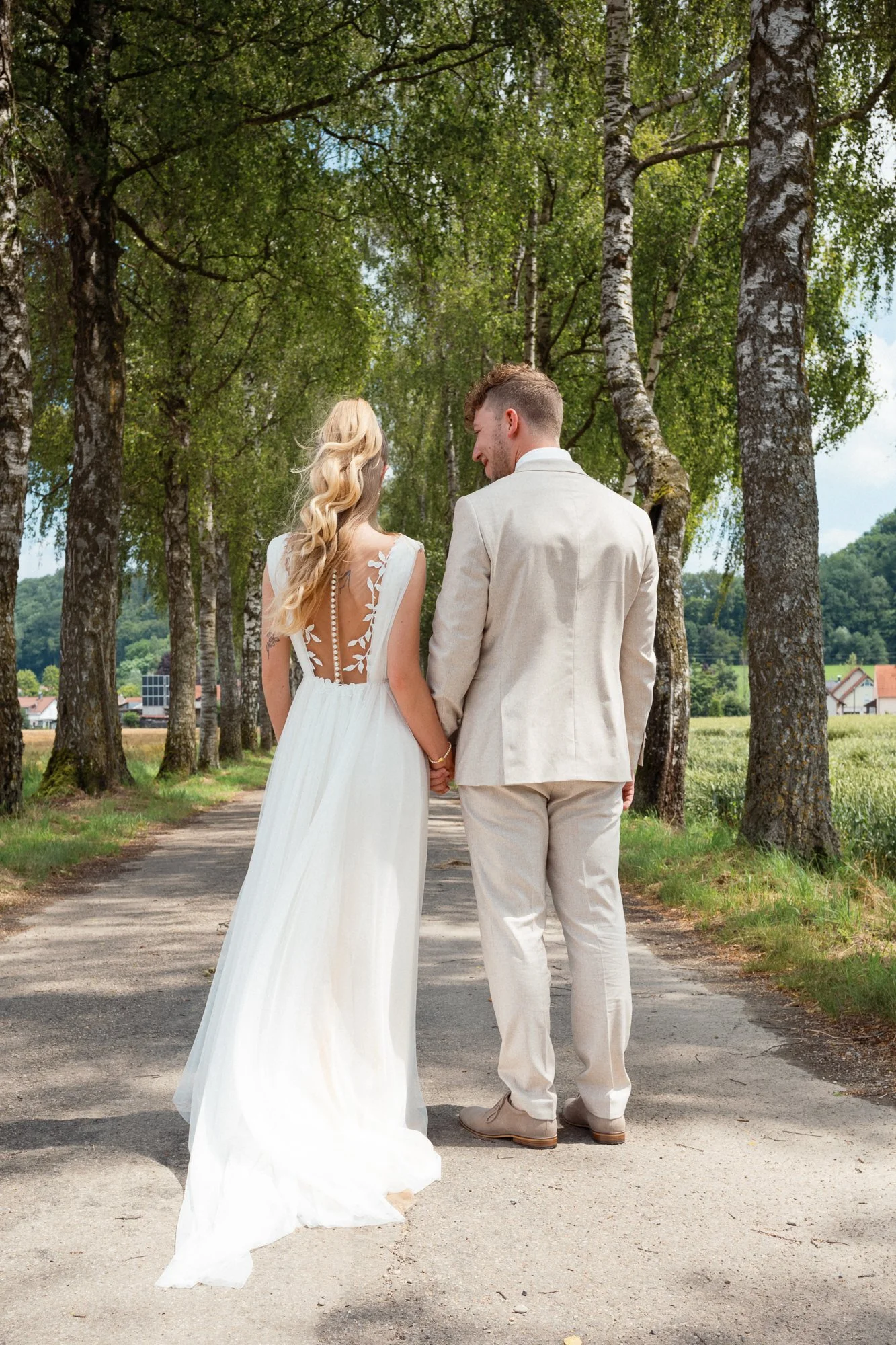Ein Brautpaar geht hand in hand auf einer Landstraße, umgeben von Bäumen, bei Sonnenschein und blauer Himmel, in einem ländlichen Gebiet mit Häusern im Hintergrund.