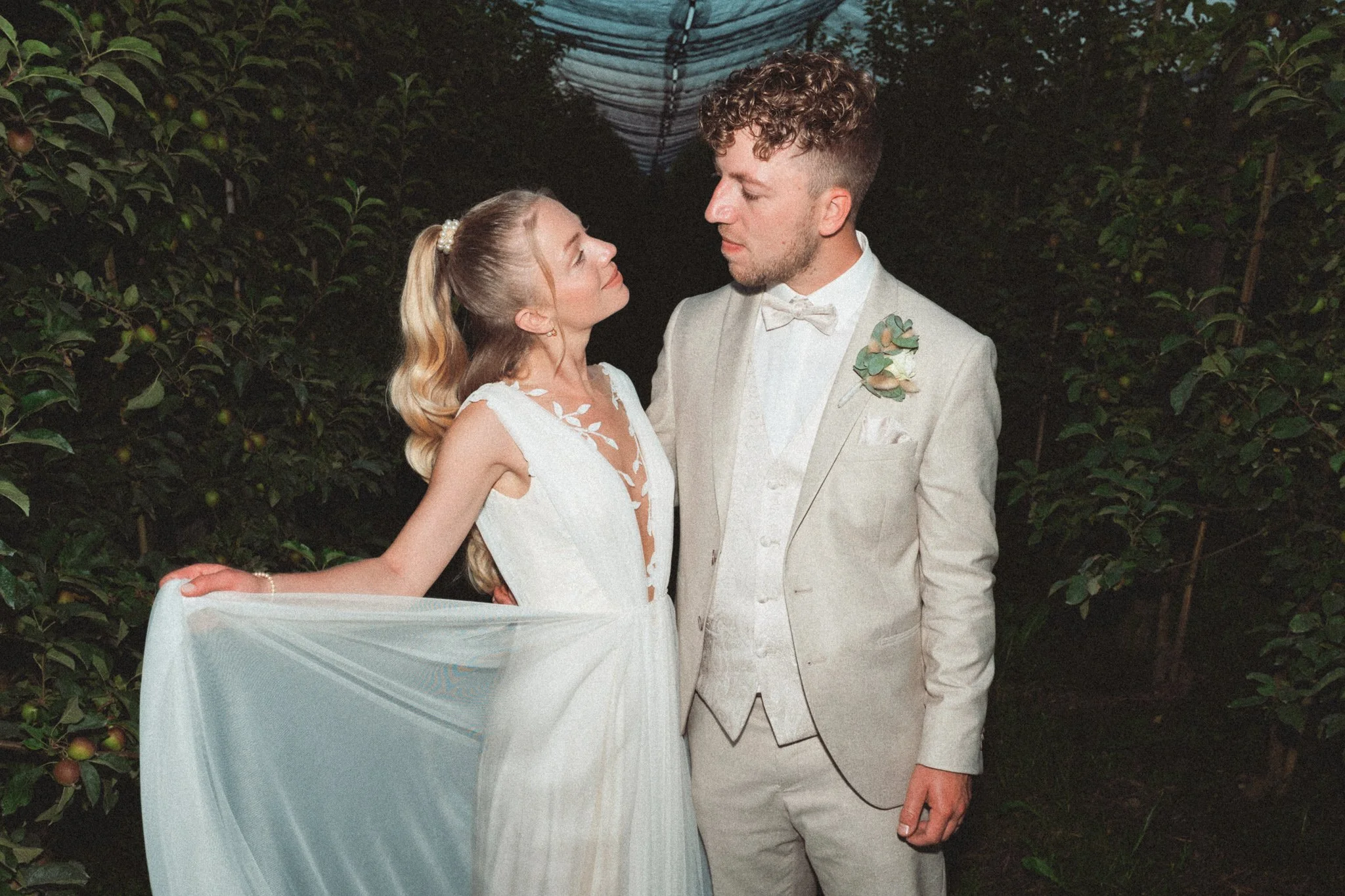 Ein frisch verheiratetes Paar bei der Hochzeit, umgeben von Bäumen, die im Hintergrund zu sehen sind, während sie liebevoll aufeinander blicken.