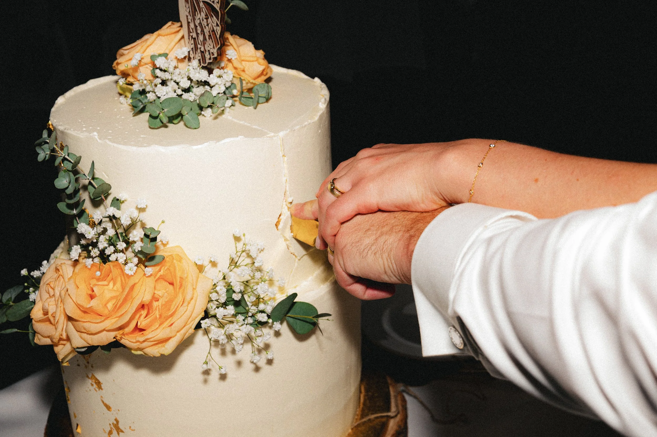 Nahaufnahme eines Hochzeitskuchens, der mit Blumen und Eukalyptus dekoriert ist, während jemand ihn anschneidet.