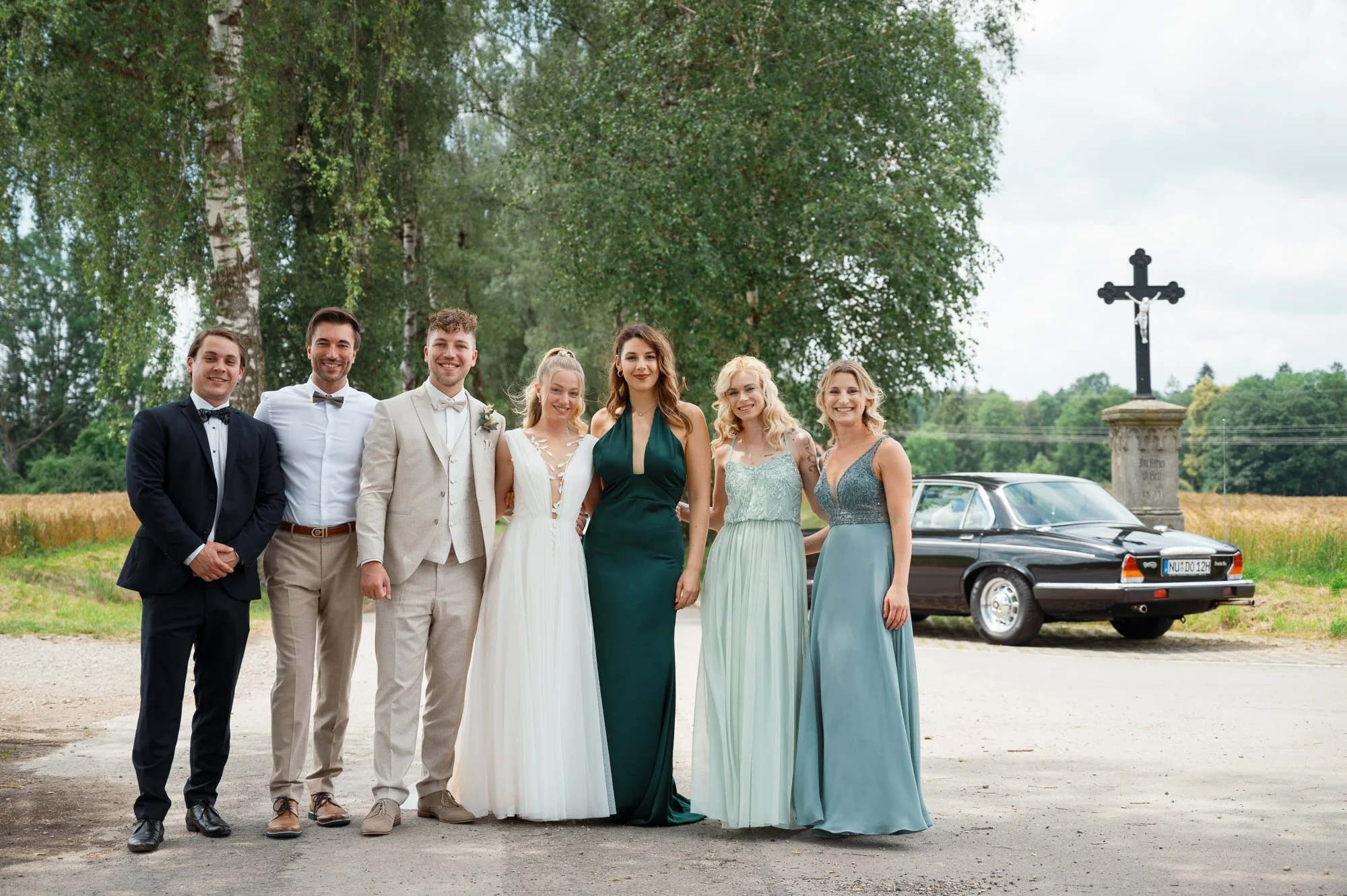 Gruppe von sechs Menschen in formeller Kleidung, vermutlich bei einer Hochzeit, im Freien mit Bäumen und einem Kreuz im Hintergrund sowie einem schwarzen Auto auf der rechten Seite.
