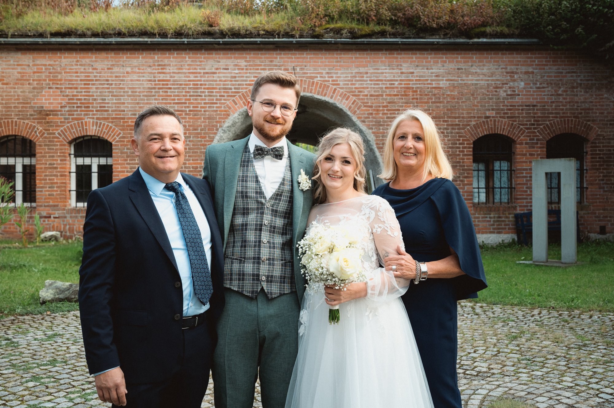 Eine Hochzeitsgruppe aus vier Personen steht vor einem roten Backsteingebäude mit großen Fenstern und einem Bogen in der Mitte. Es sind zwei Männer und zwei Frauen, darunter die Braut in einem weißen Hochzeitskleid mit Blumen im Haar, die einen Braut