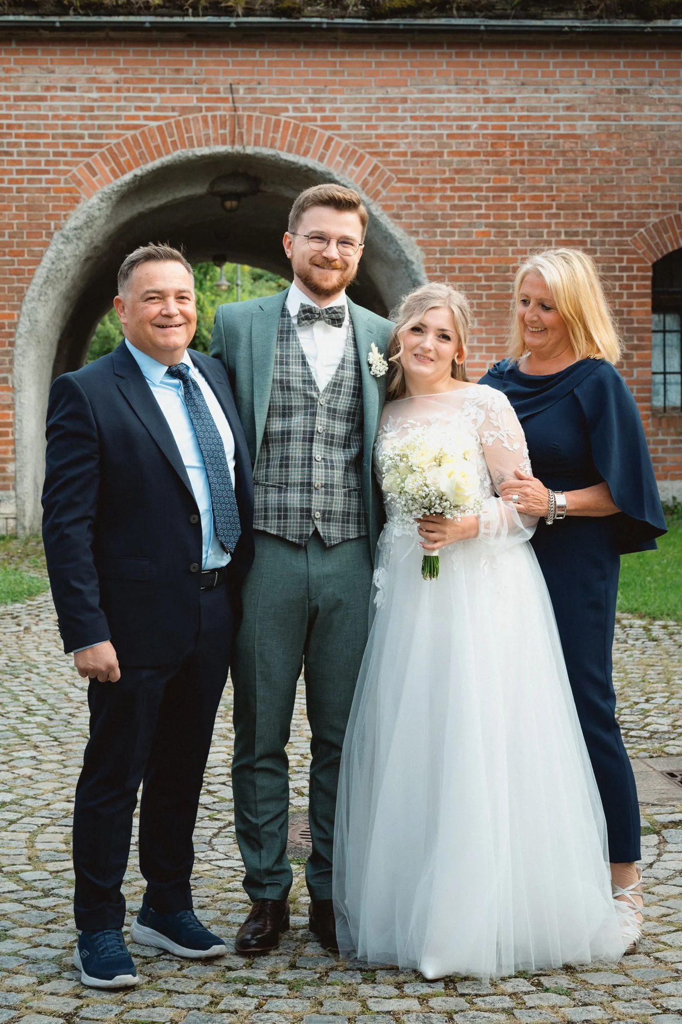 Junge Frau in Brautkleid mit Blumenstrauß, umgeben von drei Menschen bei einer Hochzeit im Freien, vor einer Backsteinmauer.