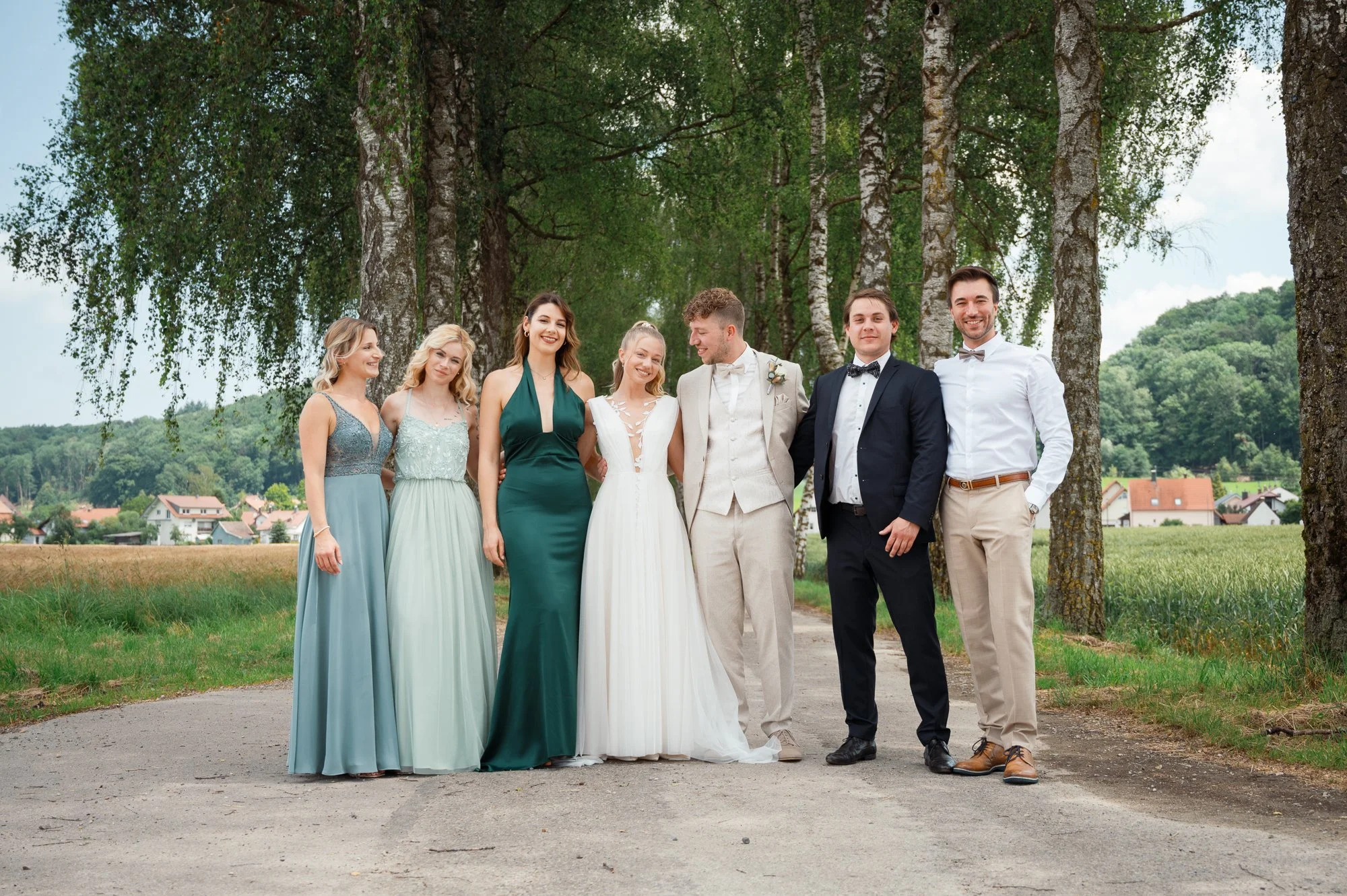 Eine Gruppe von acht Menschen, die bei einem formellen Anlass im Freien stehen, vor Bäumen und einem Dorf im Hintergrund. Die Gruppe besteht aus sechs Frauen in eleganten Kleidern und zwei Männern in Anzügen, die lachen und lässig posieren.