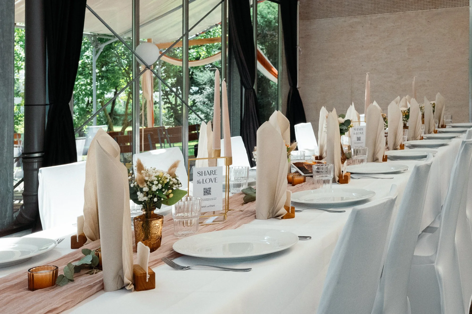 Ein festlich gedeckter langer Tisch mit weißem Tischtuch, weißen Papptellern, Stoffservietten, kleinen Blumenarrangements, Kerzen und einer kleinen Tafel mit der Aufschrift "Share the Love" in einem Raum mit großen Fenstern und Blick ins Grüne.