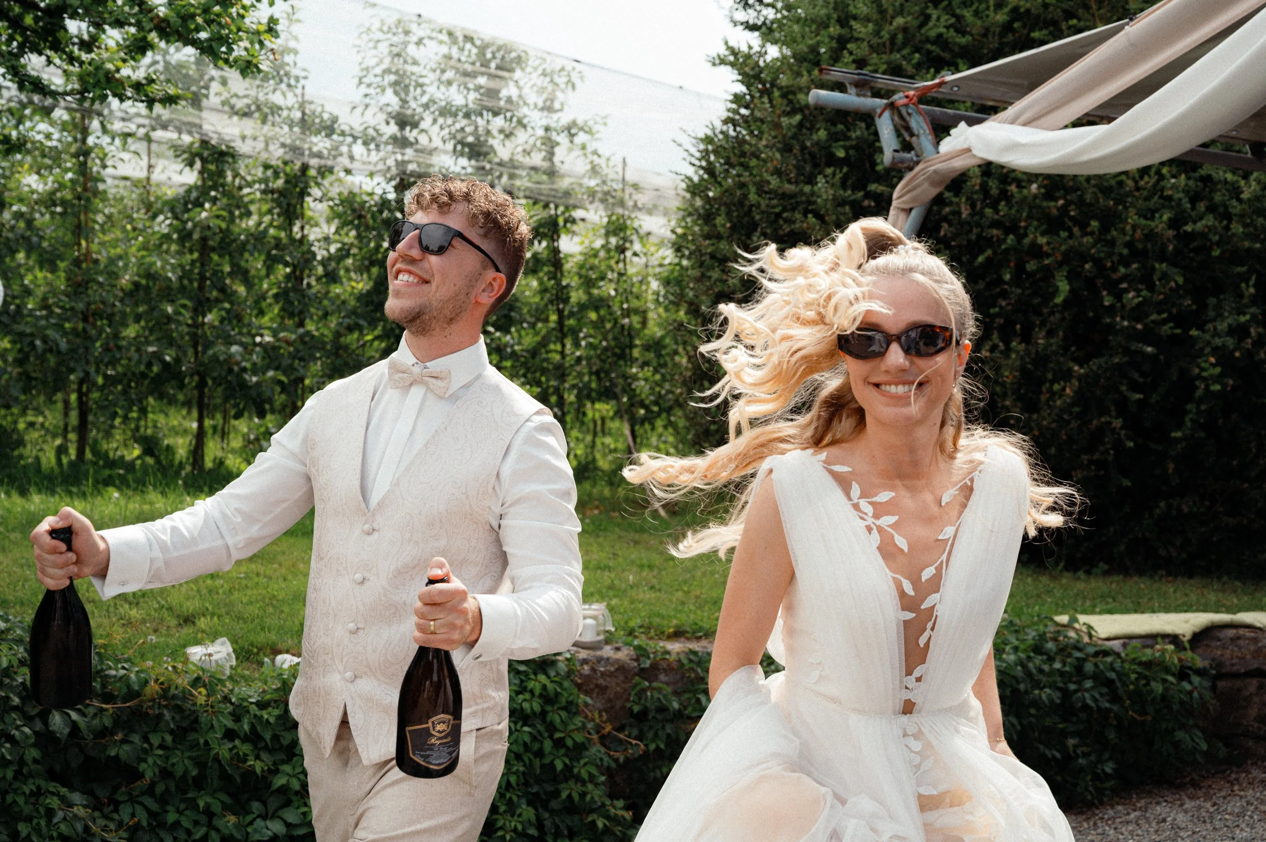 Ein Mann und eine Frau in Hochzeitskleidung, die im Garten unterwegs sind, der Mann hält zwei Sektflaschen, beide tragen Sonnenbrillen und lächeln.