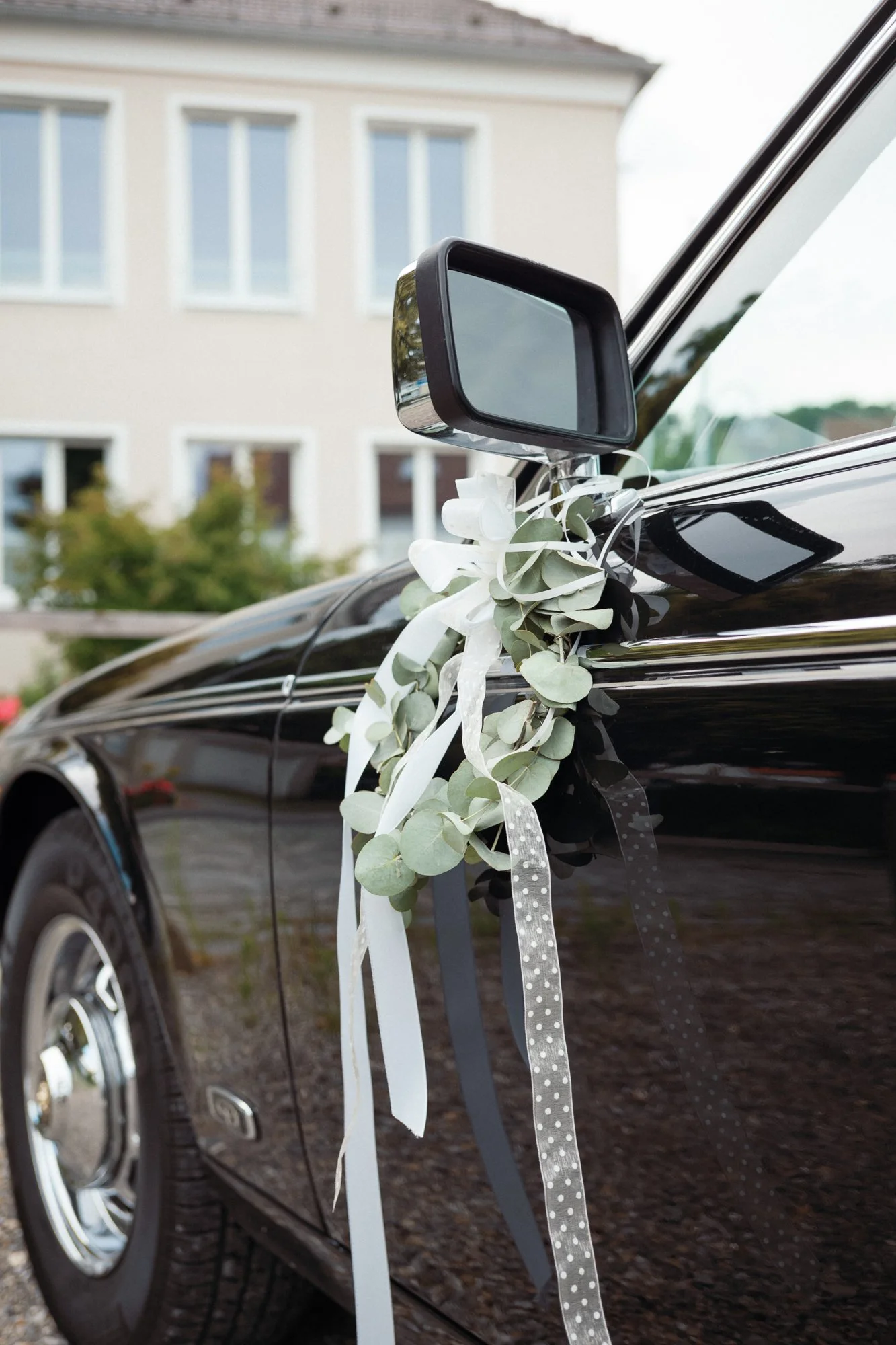 Ein schwarzes Auto mit weißer Schleife und Eukalyptuszweigen am Seitenspiegel, vermutlich für eine Hochzeit dekoriert.