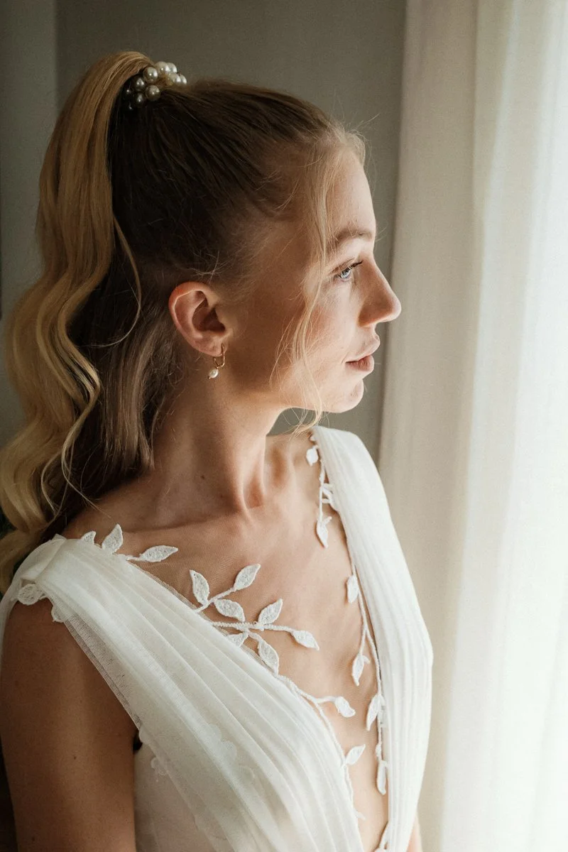 Junge Frau mit blonden Haaren, die zu einem hohen Pferdeschwanz gebunden sind, blickt durch ein Fenster. Sie trägt ein weißes Kleid mit Blättermuster an den Schultern, Ohrringe und einen dezenten Make-up-Look.