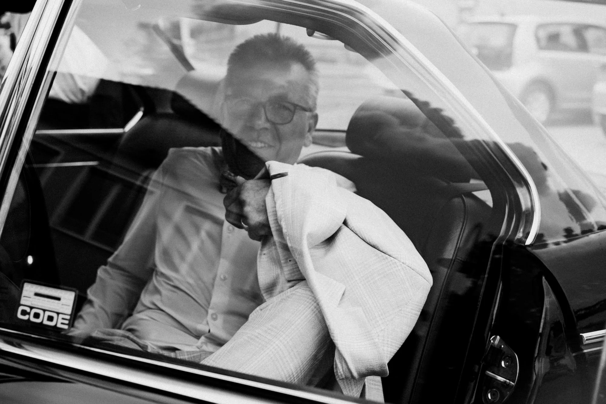 Ein Mann sitzt im Auto und trägt eine Brille und ein Armband. Er hat einen weißen Hemd an und scheint die Tür offen zu haben, während er lächelt. Das Bild ist in Schwarz-Weiß aufgenommen.