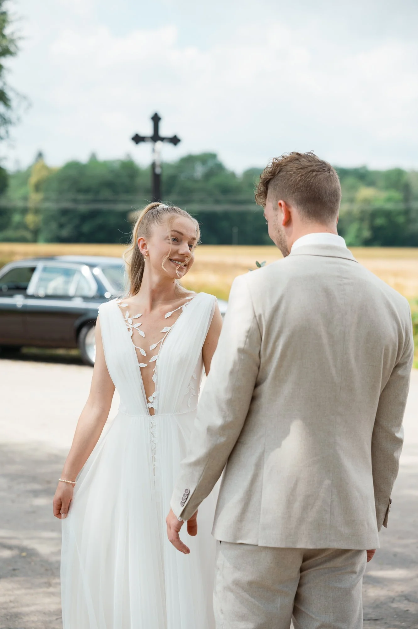 Ein Brautpaar, eine Frau in einem weißen Hochzeitskleid und ein Mann in einem hellen Anzug, hält Händchen im Freien. Im Hintergrund sind Bäume, ein Auto und ein Kreuz sichtbar.