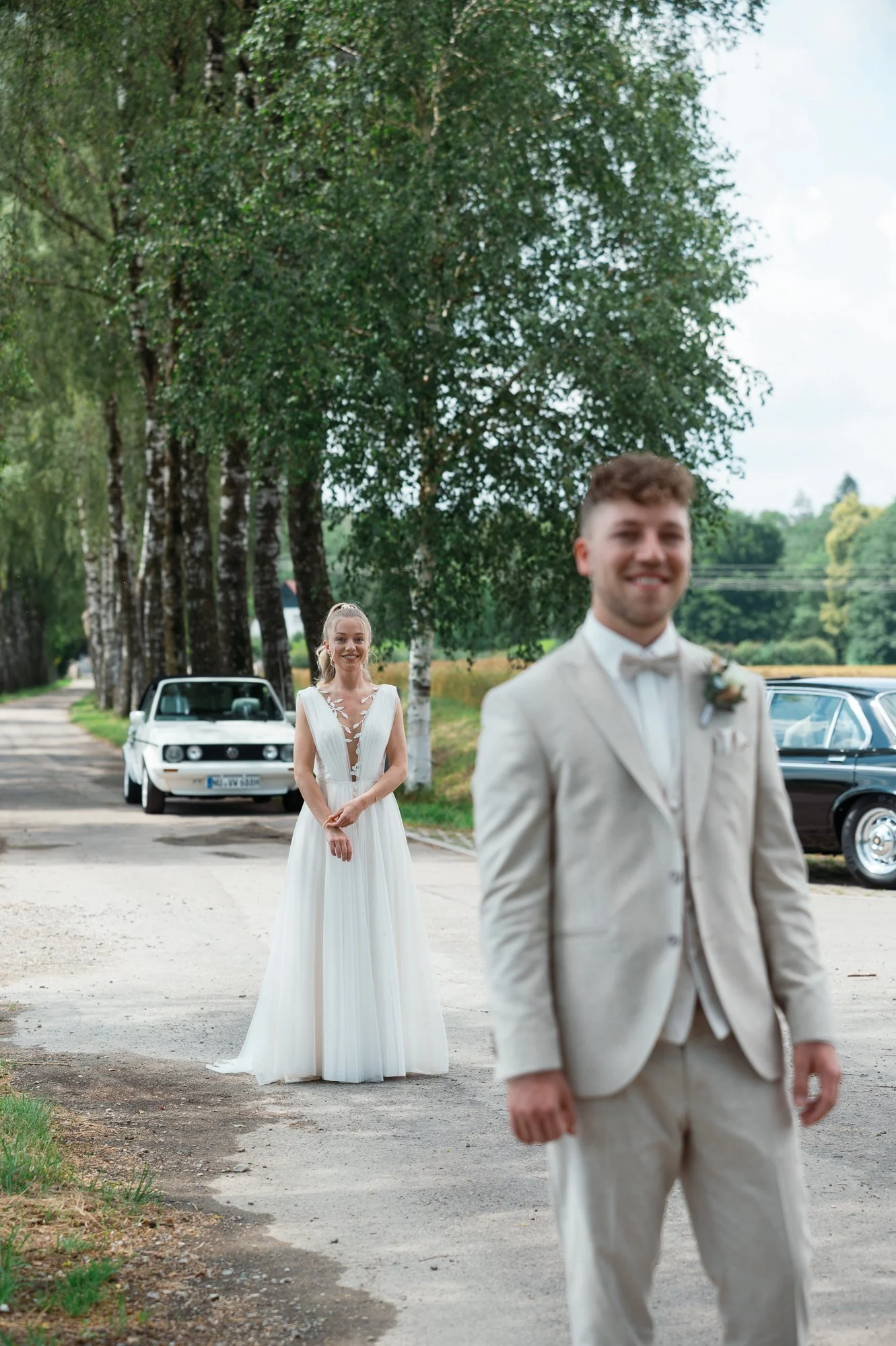 Ein Brautpaar steht auf einer Landstraße, im Hintergrund sind Bäume und alte Autos zu sehen, die Braut trägt ein weißes Hochzeitskleid, der Bräutigam einen hellen Anzug.