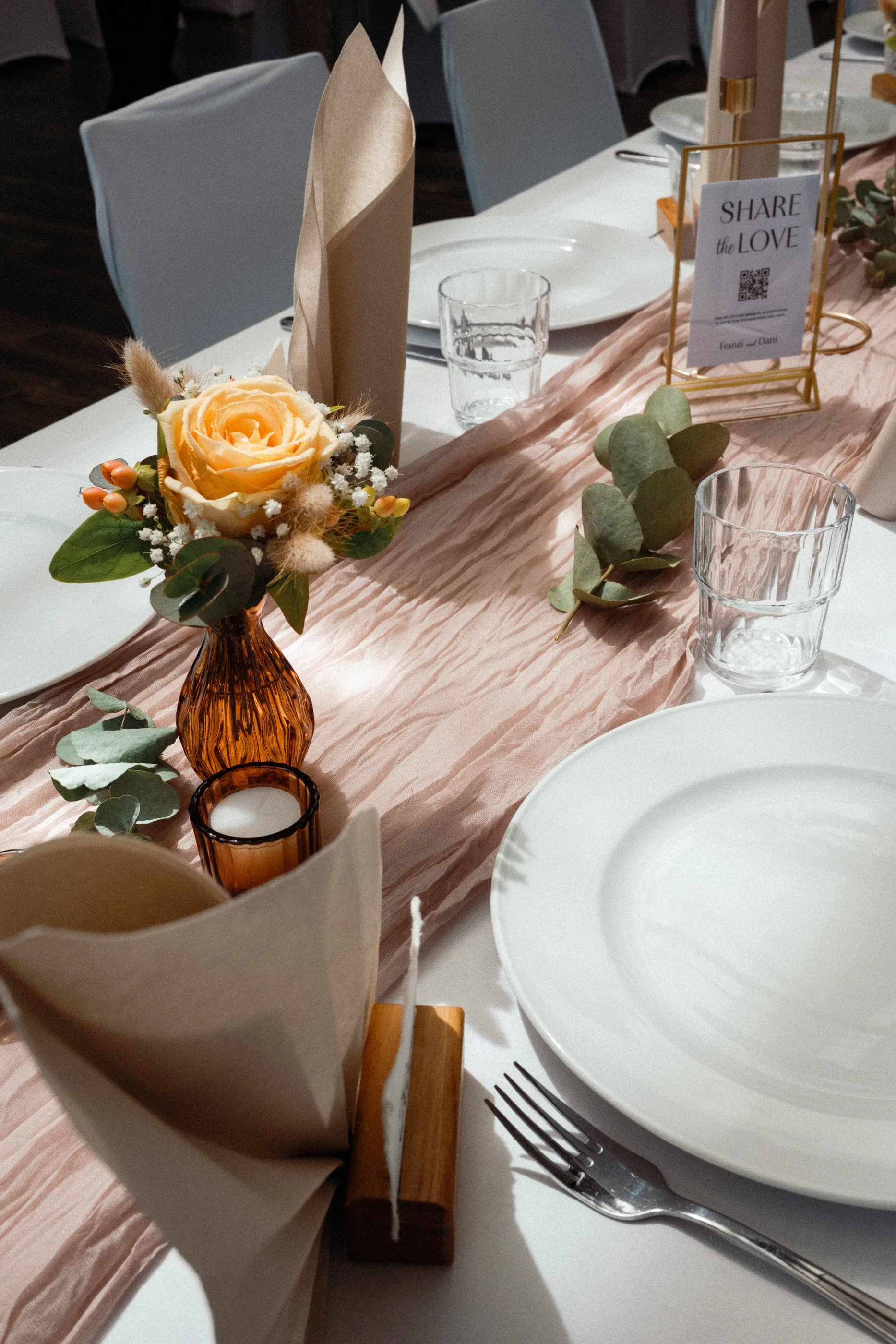 Ein gedeckter Tisch für eine Feier mit einer Blumenanordnung, Servietten, Gläsern, Tellern und Besteck, sowie eine kleine Tafel mit der Aufschrift „Share the Love“.