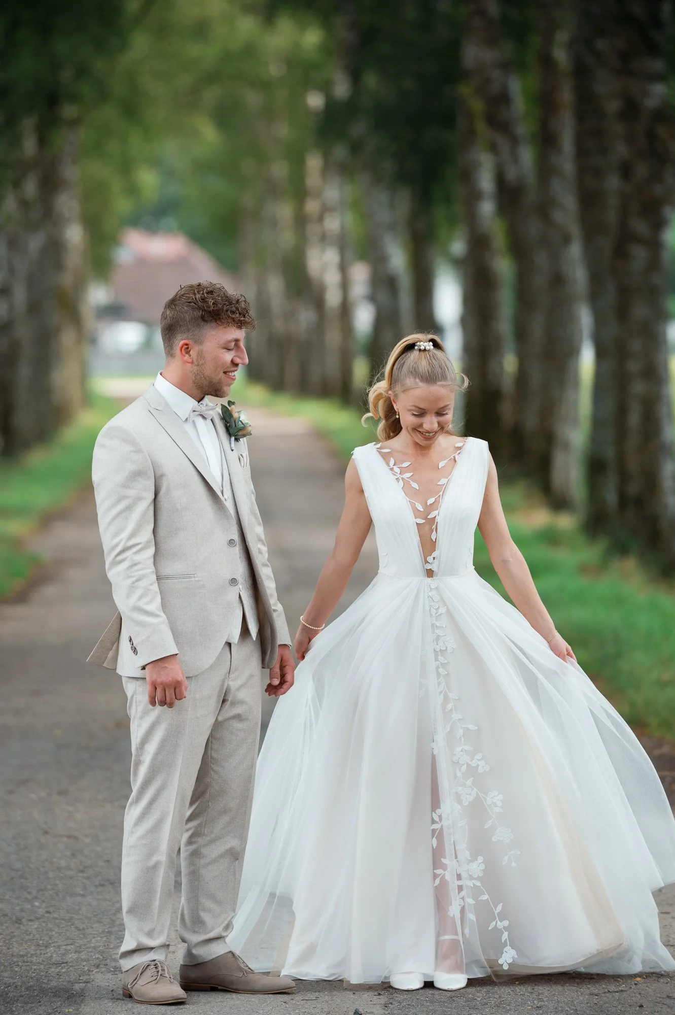 Ein Brautpaar in gelöster Atmosphäre, der Bräutigam trägt einen hellgrauen Anzug und die Braut ein weißes Kleid mit floralem Detail, sie lächelt und hält den Rock ihres Kleides hoch. Sie stehen auf einem Baumweg, umgeben von hohen Bäumen im Freien.