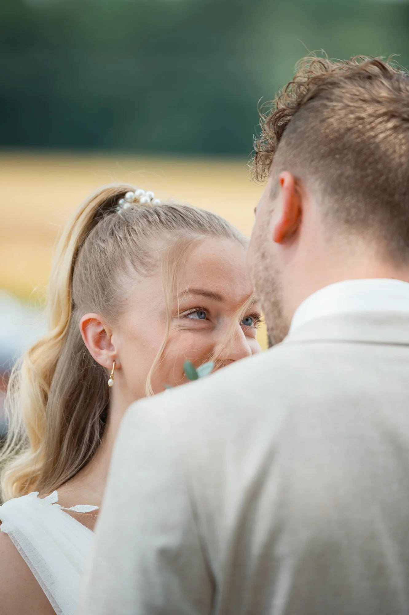 Ein Brautpaar bei einer Hochzeit. Das Mädchen hat blonde Haare mit Perlen im Haar und trägt Ohrringe. Das Paar schaut sich liebevoll an, die Szene ist im Freien mit unscharfem Hintergrund.