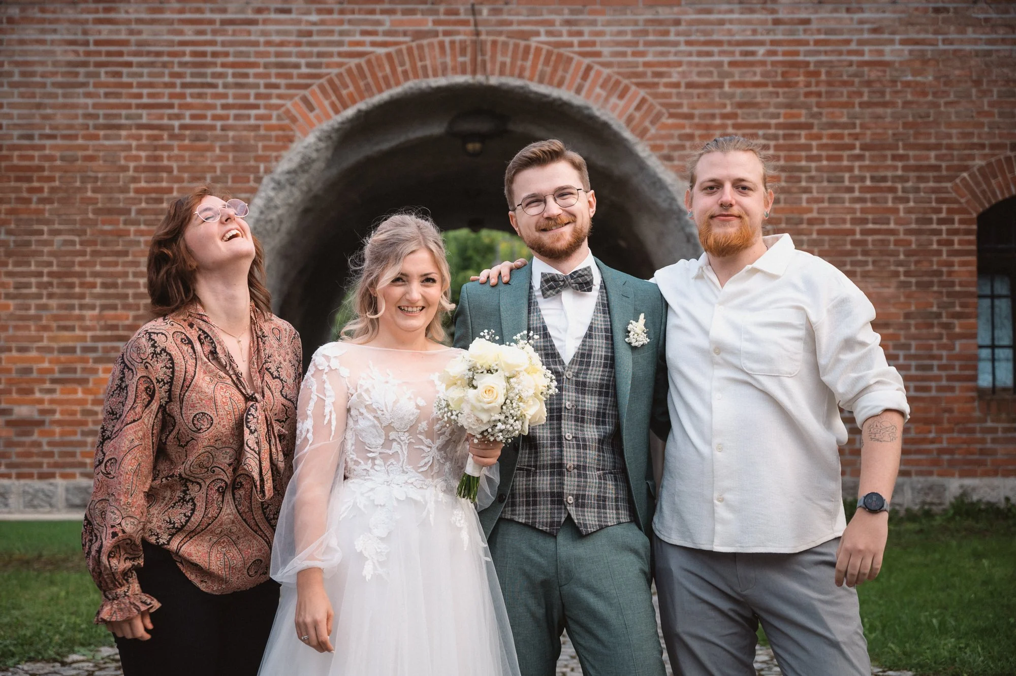 Gruppe von fünf Personen bei einer Hochzeit, sie stehen vor einer roten Backsteinmauer, lächeln und posieren für das Foto, die Braut hält einen Blumenstrauß, alle sind lässig gekleidet.
