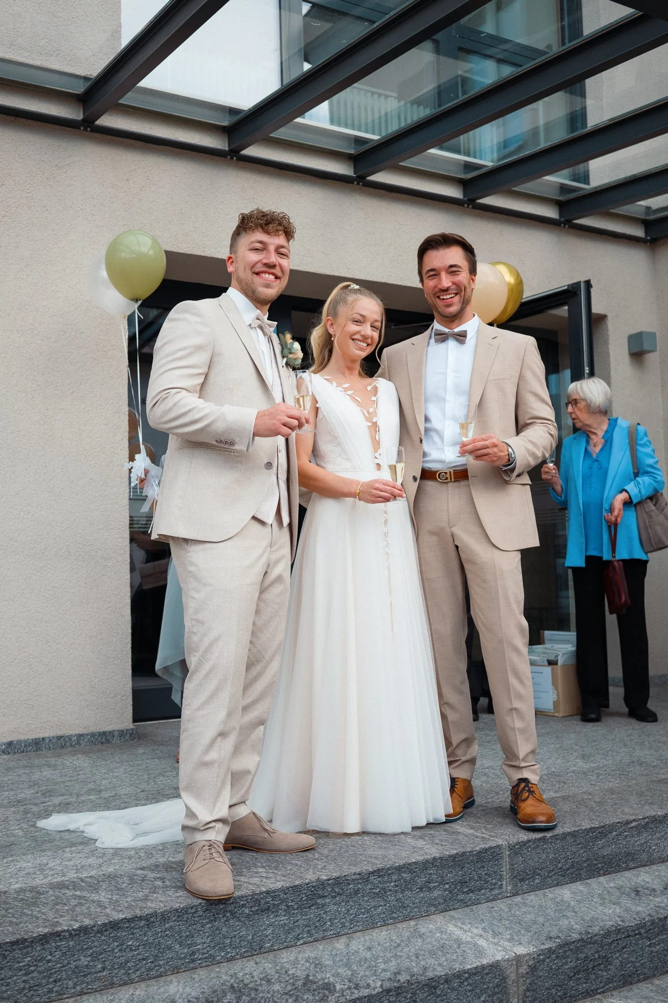 Drei Menschen bei einer Hochzeitsfeier, braut in weißem Kleid, Bräutigam und ein Freund in beige Anzügen, alle lachen und halten Gläser. Hintergrund mit einem älteren Herrn in blauer Jacke.