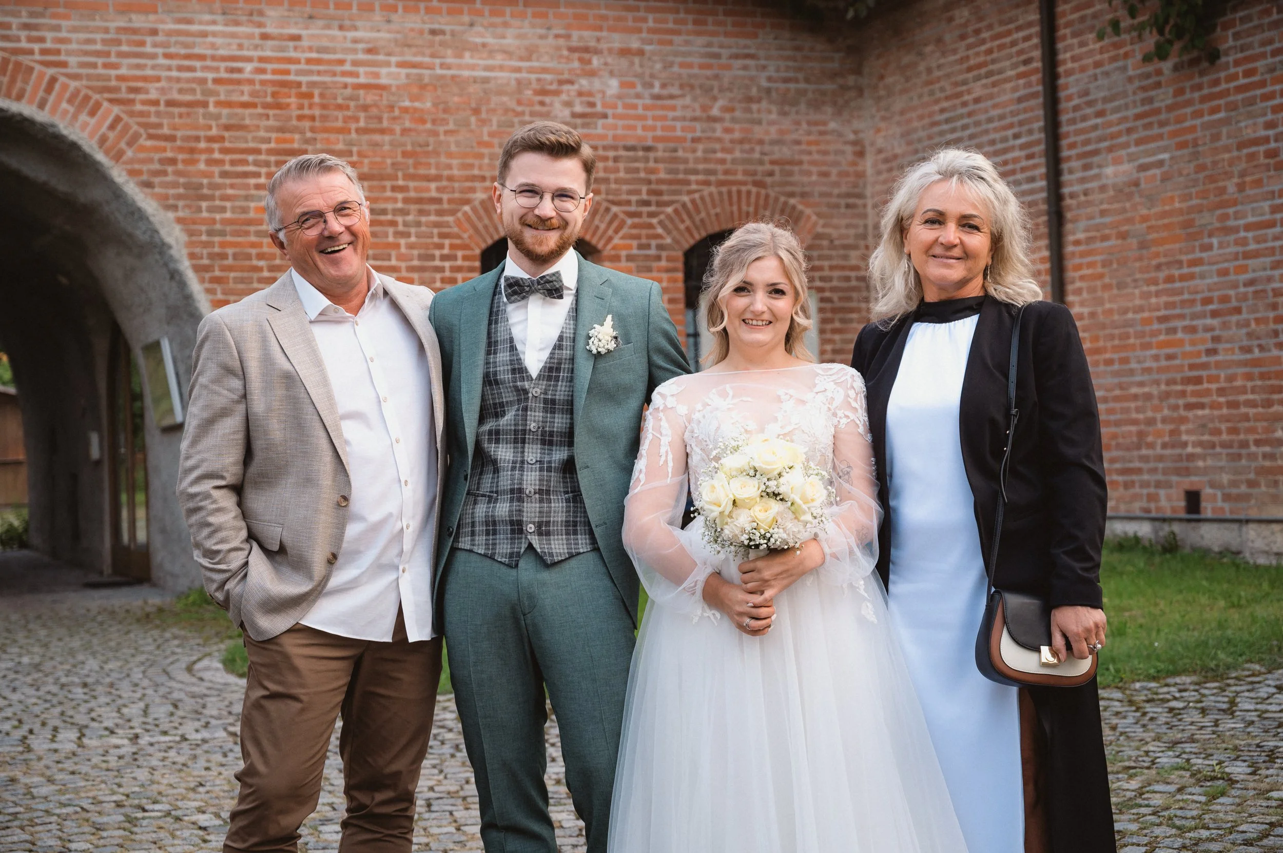 Eine Gruppe von fünf Menschen steht zusammen vor einer roten Ziegelmauer, darunter eine Braut in einem weißen Brautkleid, die ein Blumenstrauß hält, und ein Bräutigam in einem grauen Anzug mit Fliege.