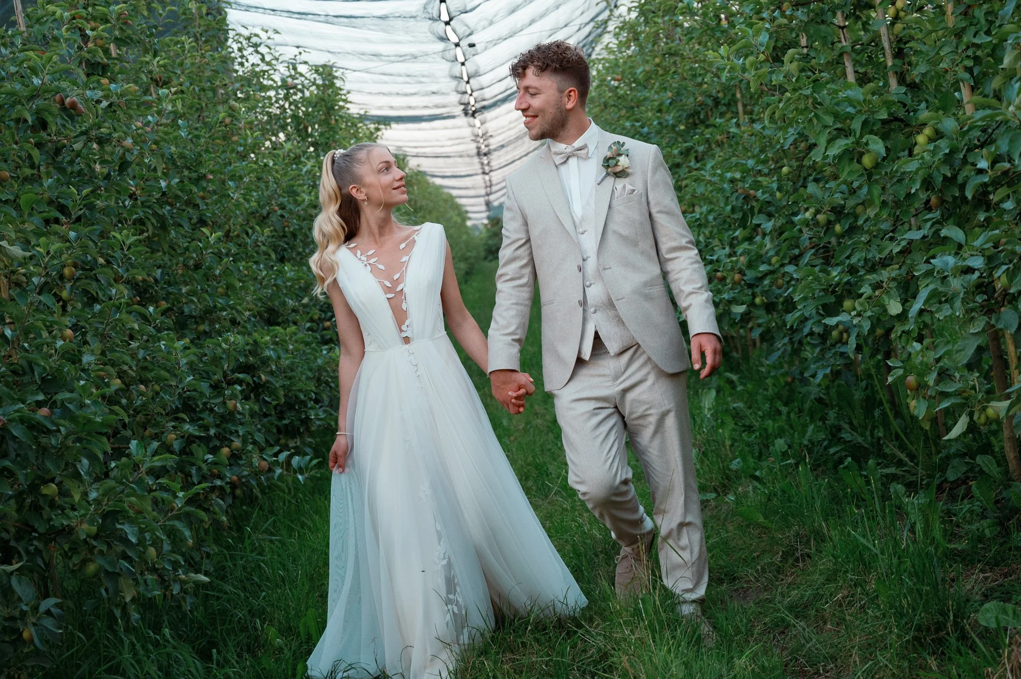 Ein glückliches Brautpaar hält Händchen in einem Obstgarten. Die Braut trägt ein weißes Brautkleid und der Bräutigam einen hellgrauen Anzug. Beide sehen sich liebevoll an.