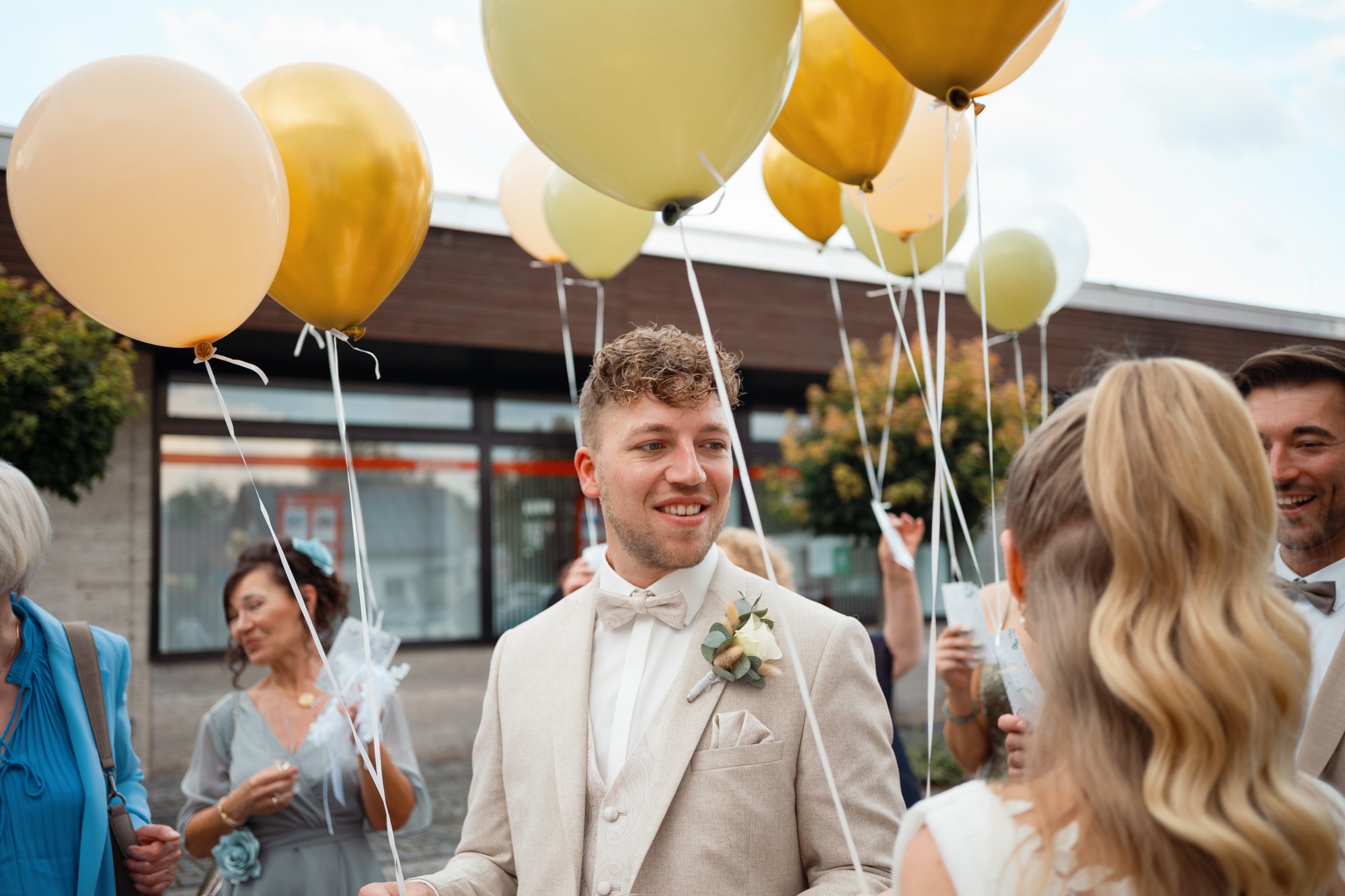 Eine Hochzeitsparty im Freien, mit Menschen, die mit Luftballons feiern, darunter ein Mann in einem hellen Anzug und Schleier. Es ist eine festliche Stimmung bei Tageslicht.