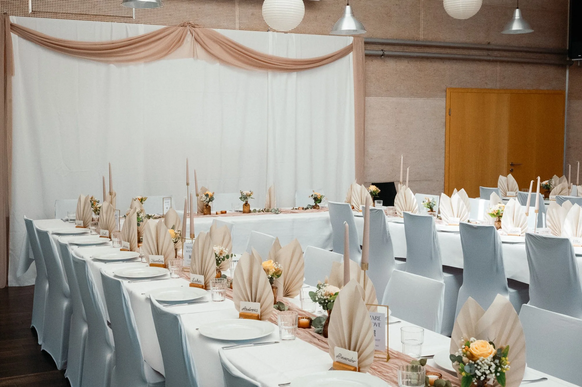 Festlich dekorierter Veranstaltungsraum mit rundem Tisch, weißen Stühlen, Kerzen, kleinen Blumengestecken, Namensschildern und Besteck, vorbereitet für eine Feier oder ein Event.