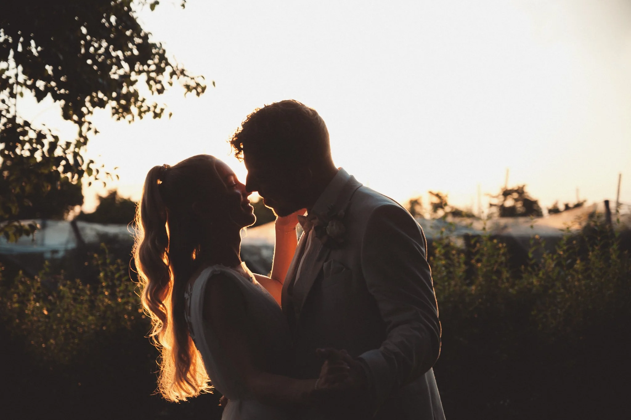 Ein Paar teilt sich einen intimen Moment im Sonnenuntergang, sie stehen sich gegenüber, ihre Stirn aneinander, im Freien mit Bäumen und Gebäuden im Hintergrund.