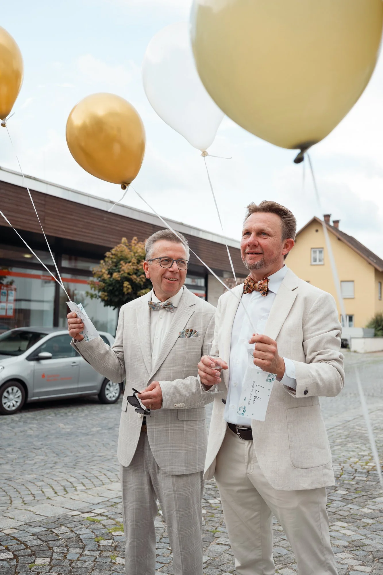 Zwei Männer in weißen Anzügen und Fliegen feiern bei einer Außenveranstaltung mit goldenen und weißen Ballons, im Hintergrund ein Gebäude und ein Auto.