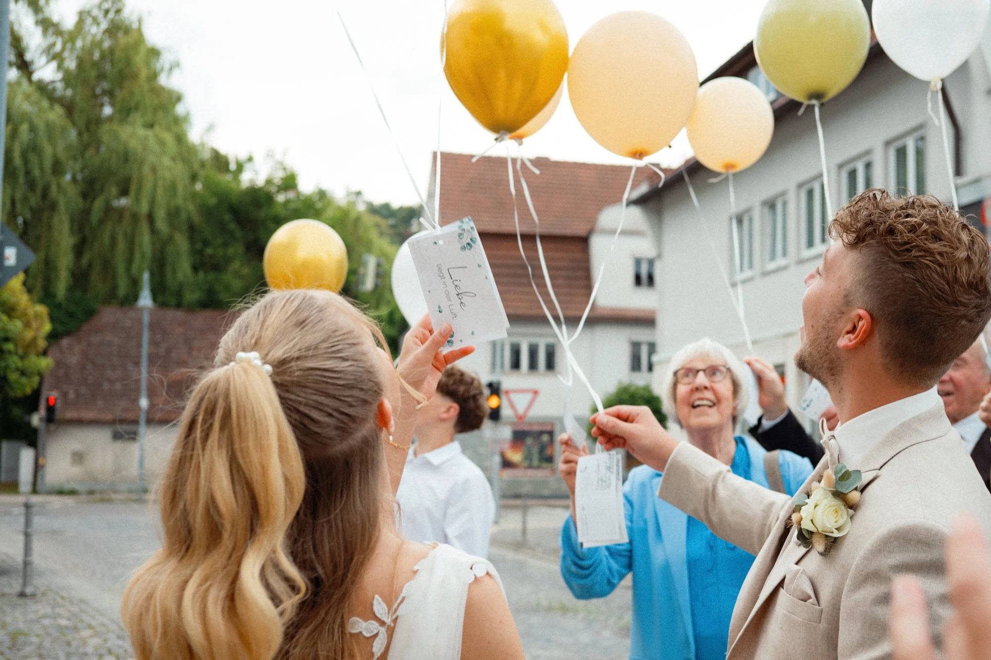 Hochzeitspaar sendet und erhält Ballons bei einer Feierlichkeit auf der Straße, umgeben von Gästen und einer Stadt im Hintergrund.
