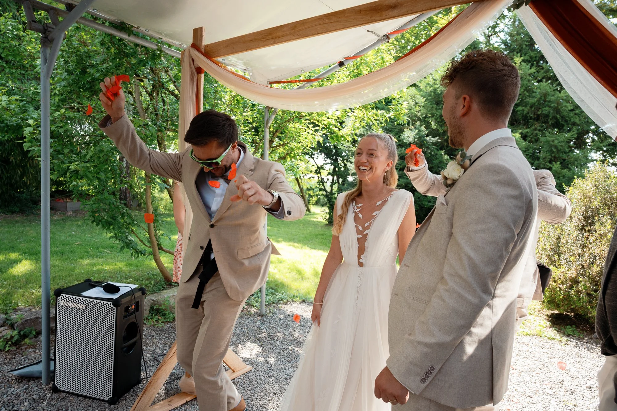 Hochzeitspaar mit Brautstrauß im Garten, umgeben von Gästen, unter einem Zelt mit Dekorationen.