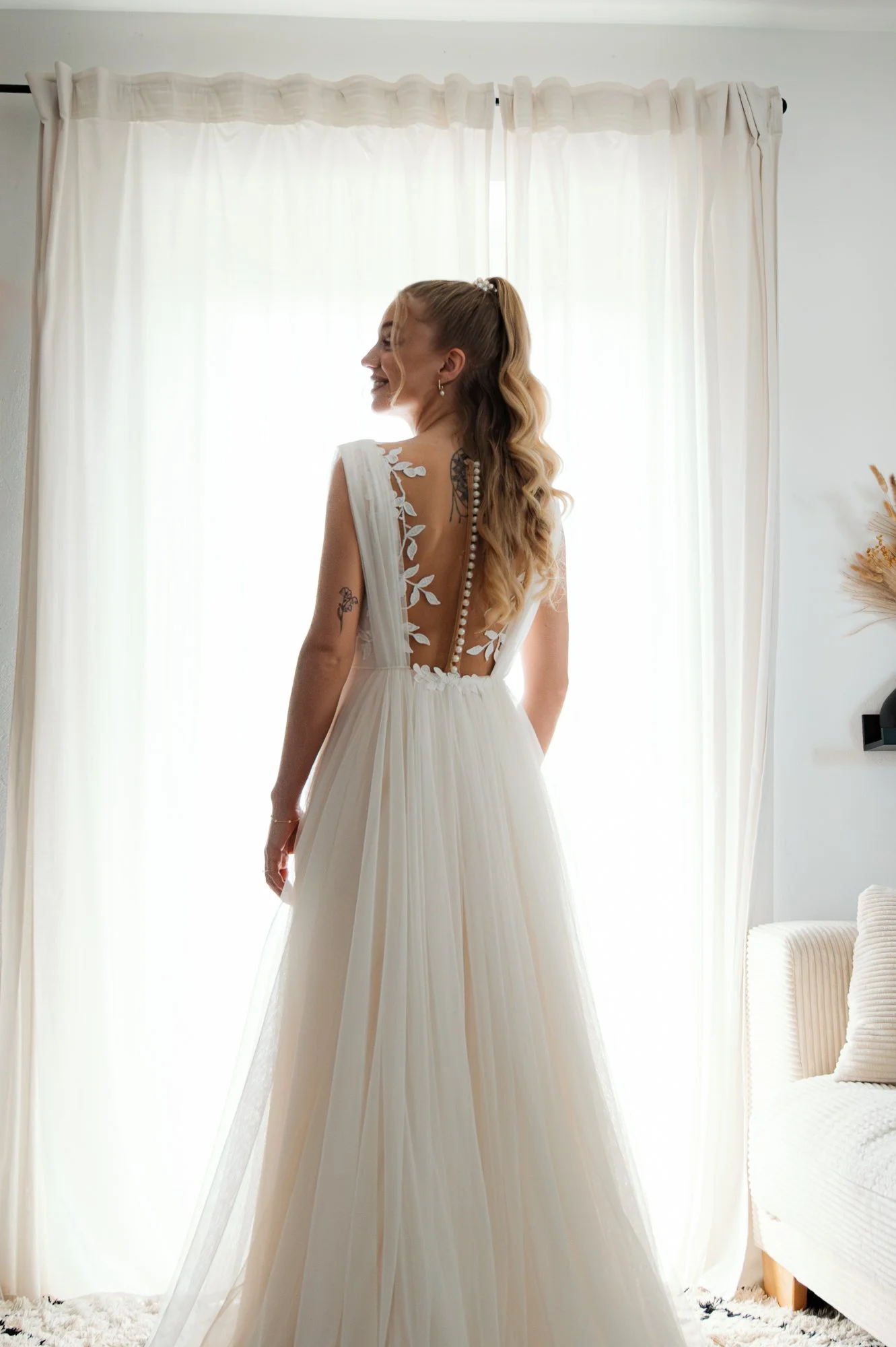 Eine lächelnde Frau im weißen Brautkleid mit offenen, gelockten Haaren vor einem weißen Vorhang mit natürlichem Licht. Das Kleid hat eine offene Rückenpartie mit Stickerei und eine Reihe von Perlen. Die Szene ist hell und ruhig, mit einem Sofa im Hin