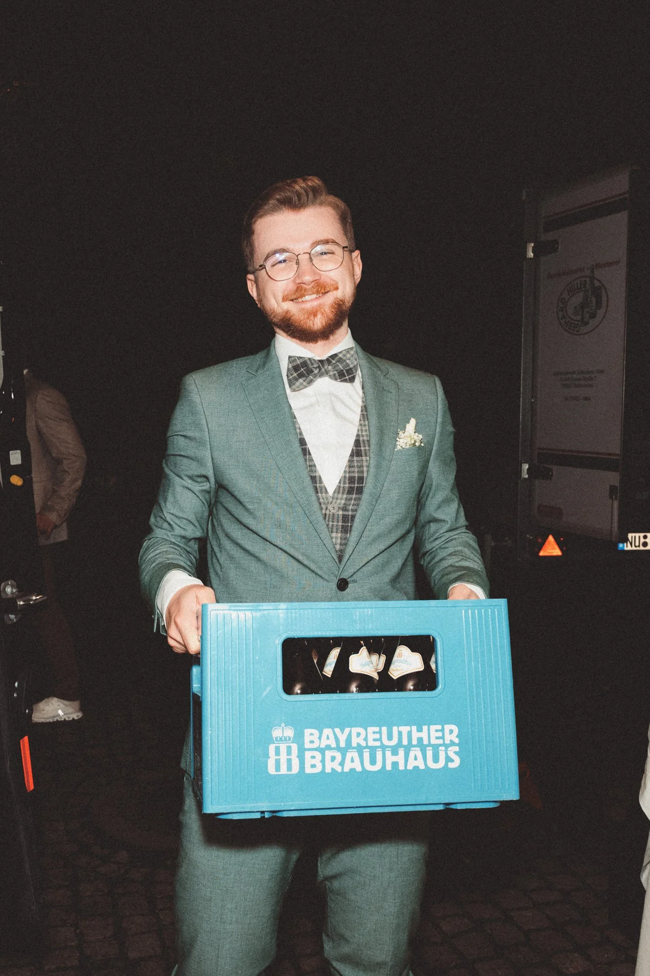 Ein lachender Mann in einem grünen Anzug mit einer Fliege hält eine blaue Kiste mit Bierflaschen, die mit dem Logo des Bayreuther Brauhauses bedruckt ist.