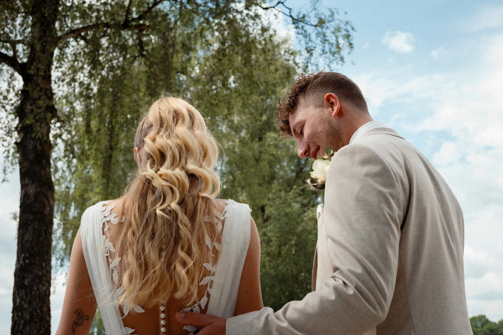 Ein Brautpaar steht im Freien unter Bäumen. Die Braut hat lange, blonde, gewellte Haare und trägt ein weißes Kleid mit Blumenmuster. Der Bräutigam trägt einen hellen Anzug und hält die Braut an der Hand. Es ist ein sonniger Tag mit blauem Himmel.