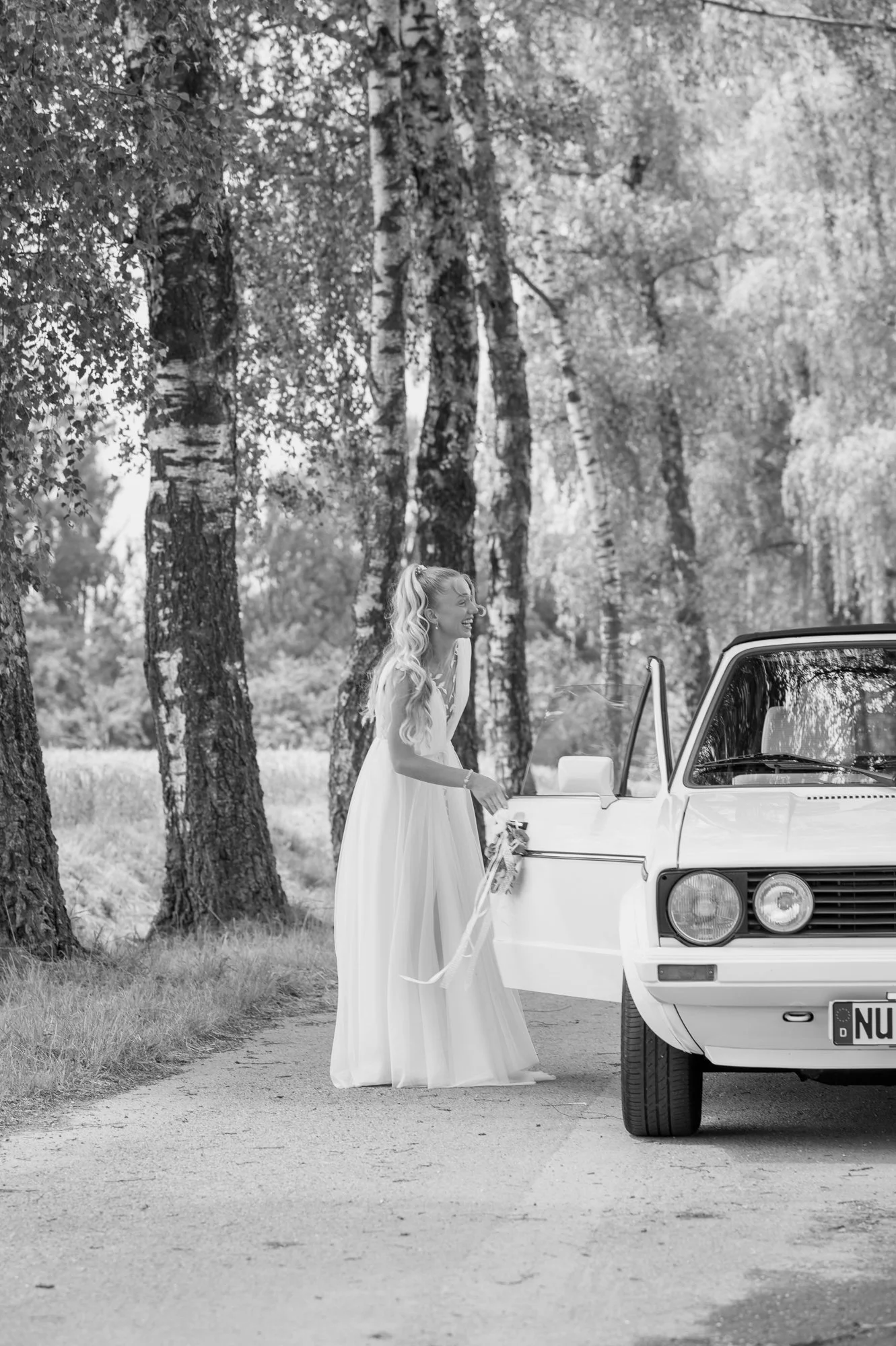Braut in weißem Kleid steht neben einem weißen Auto im Wald, lächelt und hält eine Rose.